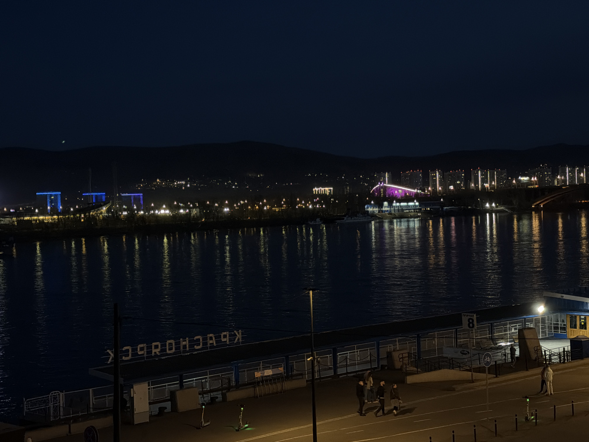 This nocturnal image captures a panoramic view of the Krasnoyarsk waterfront in Russia. The scene is dominated by a wide body of water, likely the Yenisei River, which occupies the middle ground and reflects the vibrant city lights from the far bank. Across the river, the cityscape glows with numerous illuminated buildings, including prominent blue-lit structures on the left and a large, distinctly purple-lit building, possibly a stadium or major architectural landmark, towards the right. A dark, hilly or mountainous landscape forms the distant backdrop under a deep blue night sky.
In the foreground, a paved promenade or dock area runs parallel to the water. Several people are visible walking along this path, illuminated by overhead streetlights. Two electric scooters are parked on the side. Along the edge of the dock, large white letters spell "КРАСНОЯРСК" (Krasnoyarsk), visible in reverse from this perspective, confirming the city. A sign further down the promenade reads "ПРИЧАЛ 8" (Pier 8) and displays a bicycle symbol. The time is deep night, characterized by artificial lighting, a clear sky, and the calm, reflective surface of the water. The overall atmosphere is peaceful and urban.