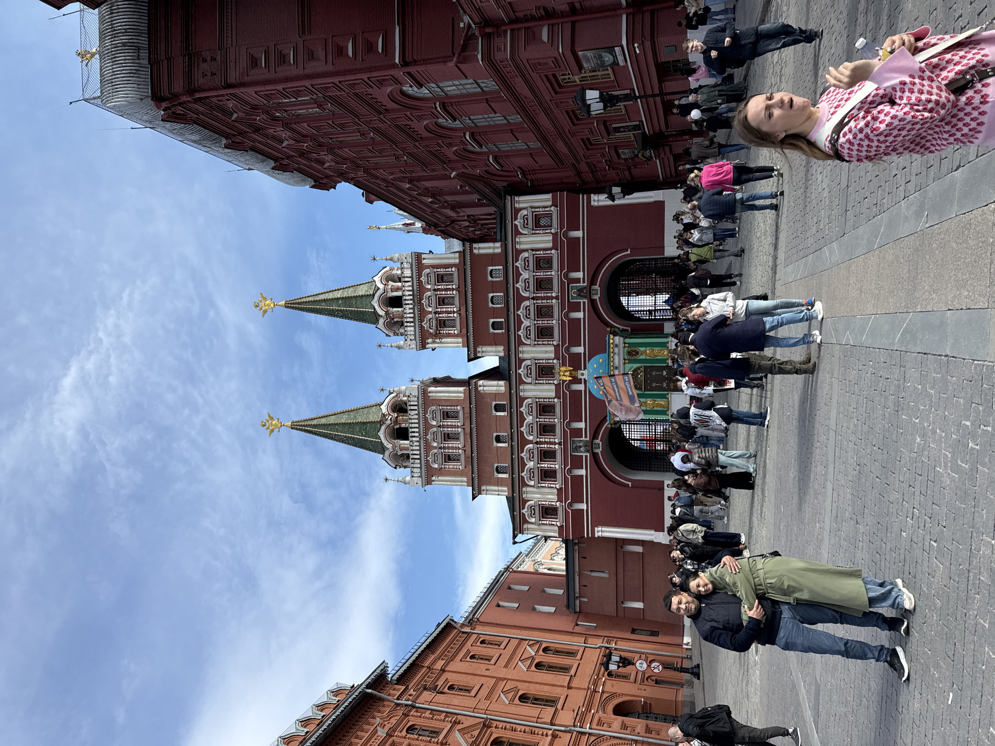 The image shows a view of Red Square in Moscow, Russia. The Resurrection Gate and the State Historical Museum are visible. There is a crowd of people walking around the square. A woman in a pink patterned sweater stands on the right side of the frame, and a couple poses near the bottom. The sky is blue with scattered clouds.