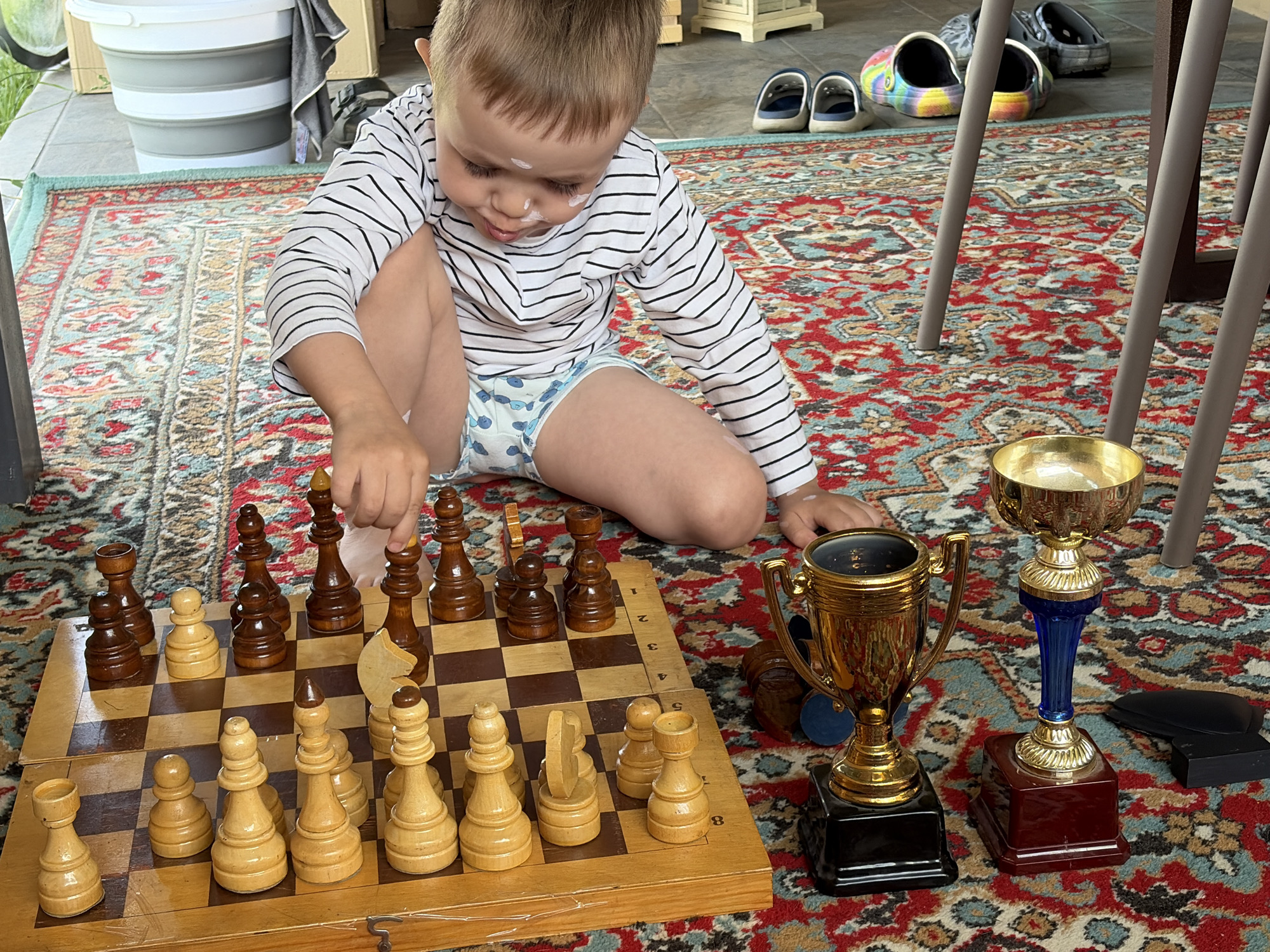 A young boy is sitting cross-legged on an oriental rug, playing chess. The chessboard is positioned in front of him. He is wearing a long-sleeved, striped shirt and a diaper. Next to the chess board are three trophies. In the background, there is a white bucket, a pair of shoes, and chair legs. The location is likely in a home, and based on the file name, it is likely in the city of Chaikovsky, Russia.