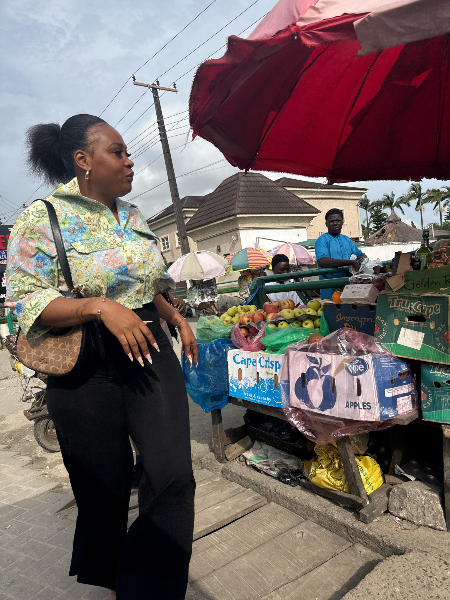 In Ado, Nigeria, a woman with her hair in a high ponytail stands at a street market stall. She is wearing a colorful, floral-patterned cropped shirt and black flared pants. A brown crossbody bag with a monogram pattern hangs from her left shoulder. She is looking to her left, with her right hand resting on her hip. The stall is laden with produce, including crates of apples and oranges. A red umbrella shades part of the stall. In the background, there are buildings and palm trees, suggesting a tropical urban environment. Two men are visible behind the stall, one in a blue shirt and the other in a white shirt. A power pole with electrical wires is also present in the scene. The ground is paved with grey rectangular tiles.