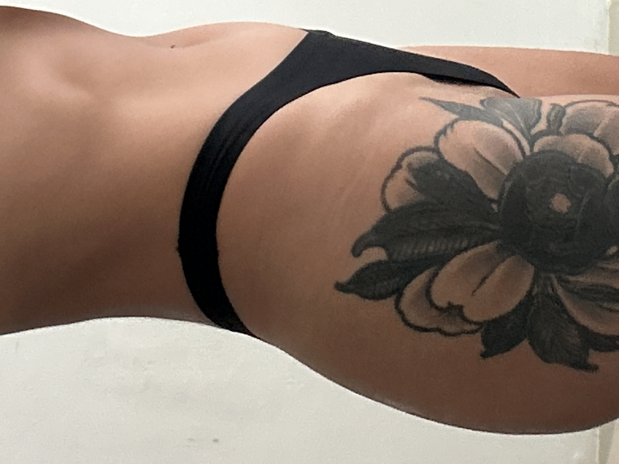 The image is a close-up, profile view of a person's hip and thigh, with a portion of their torso visible at the top. The person has a tattoo of a large flower on their thigh. They are wearing black underwear. The background is a plain white wall. The lighting suggests an indoor environment. The image is cropped tightly, focusing on the tattoo and the contour of the body.