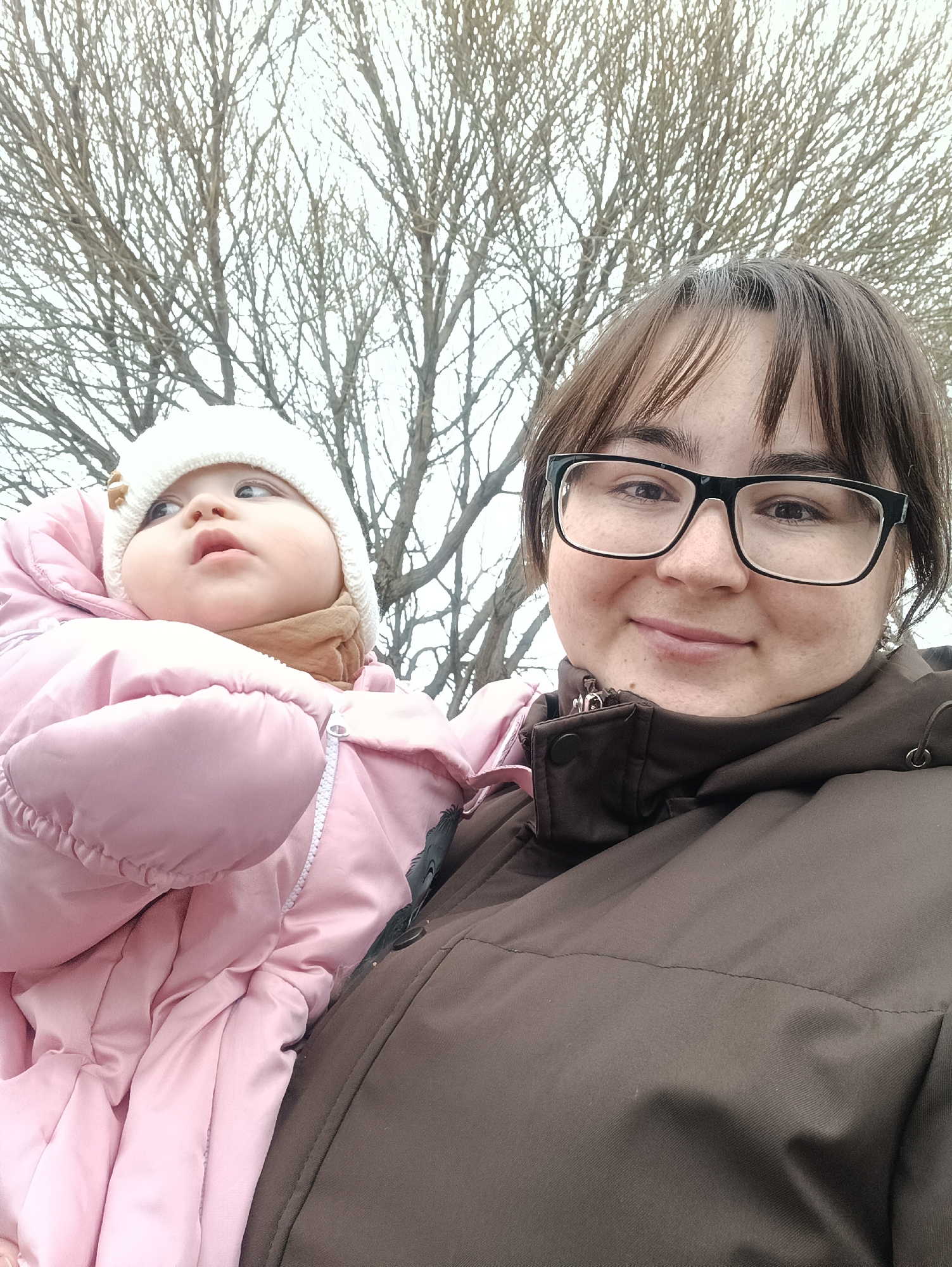 A woman holds a baby in front of a tree with bare branches. The woman is wearing glasses and a brown jacket. The baby is wearing a pink snowsuit and a white hat. The location appears to be Chaikovsky, Russia.