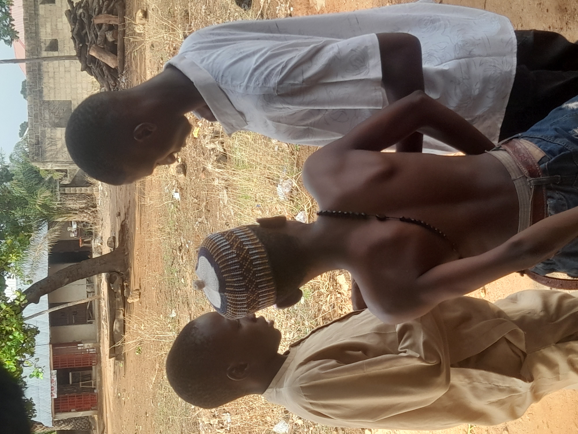 The image appears to be taken in Akwanga, Nigeria. The foreground features three young African males, two fully clothed and one shirtless wearing a blue denim jacket, light-colored undershorts, and a brown belt. One is wearing a white shirt and the other a tan garment. The middle male has a knit cap and a beaded necklace. They are outdoors in a dusty area. The background contains buildings of cinder block construction and piles of wood.