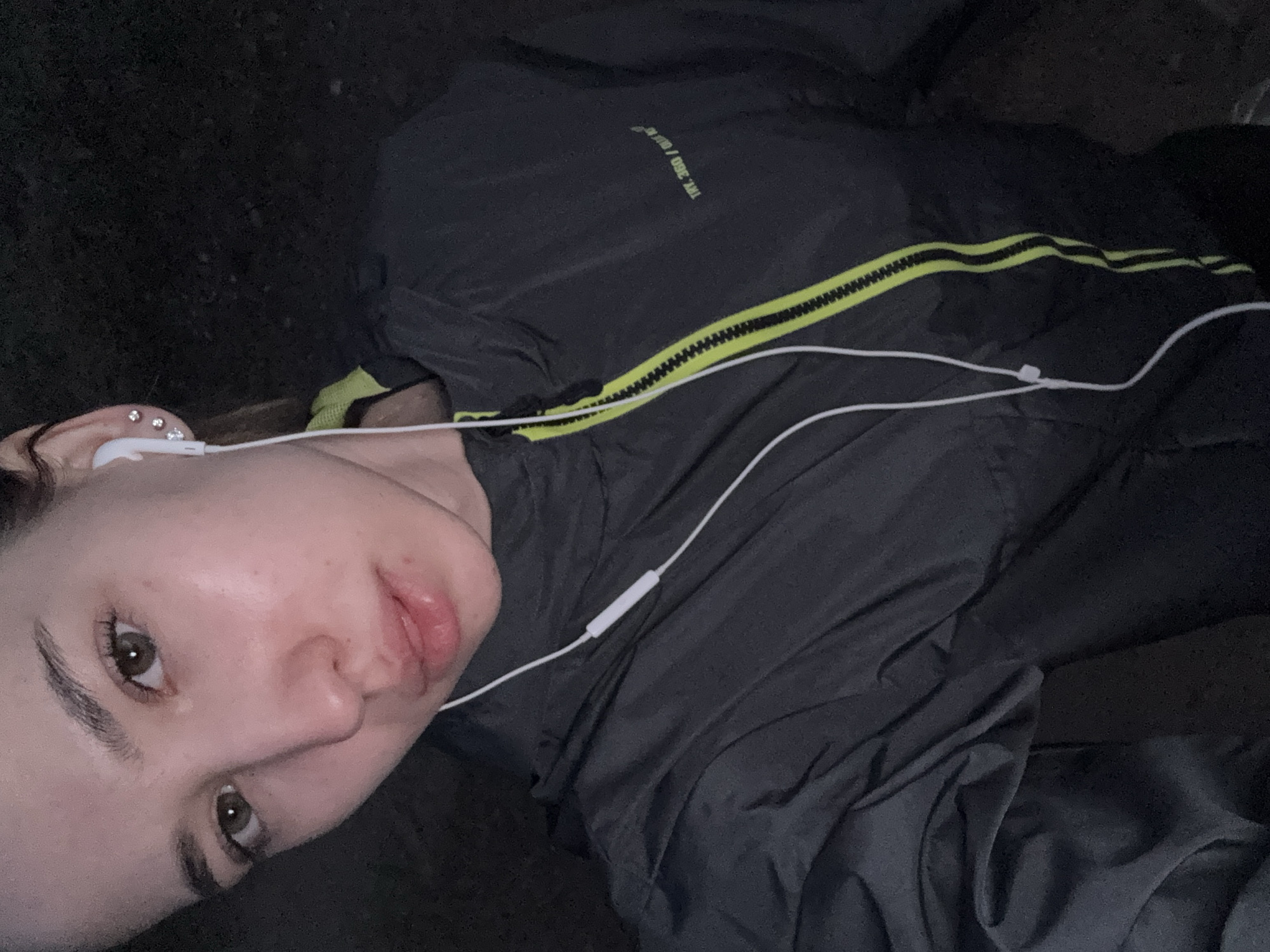 A close-up, selfie-style portrait depicts a young woman in Noviy, Russia, captured in a dimly lit environment, likely at night. She is positioned on her side, looking towards the camera with a calm, neutral expression. Her eyes are brown, and she has dark, well-defined eyebrows. Her skin appears smooth and slightly reflective.
In her visible ear, she wears multiple small, sparkling stud earrings, and a white wired earbud is placed, with its cable extending across her chest. She is dressed in a dark gray or black jacket, featuring a bright yellow-green zipper that runs diagonally. Below the zipper, two thin, horizontal yellow-green stripes are visible. On the upper part of the jacket, rotated, the text "THR. 360 / OUR" can be seen. The background is uniformly dark and indistinct, providing no specific location details beyond the low-light conditions. The woman appears to be engaged in listening, possibly to music or a podcast.