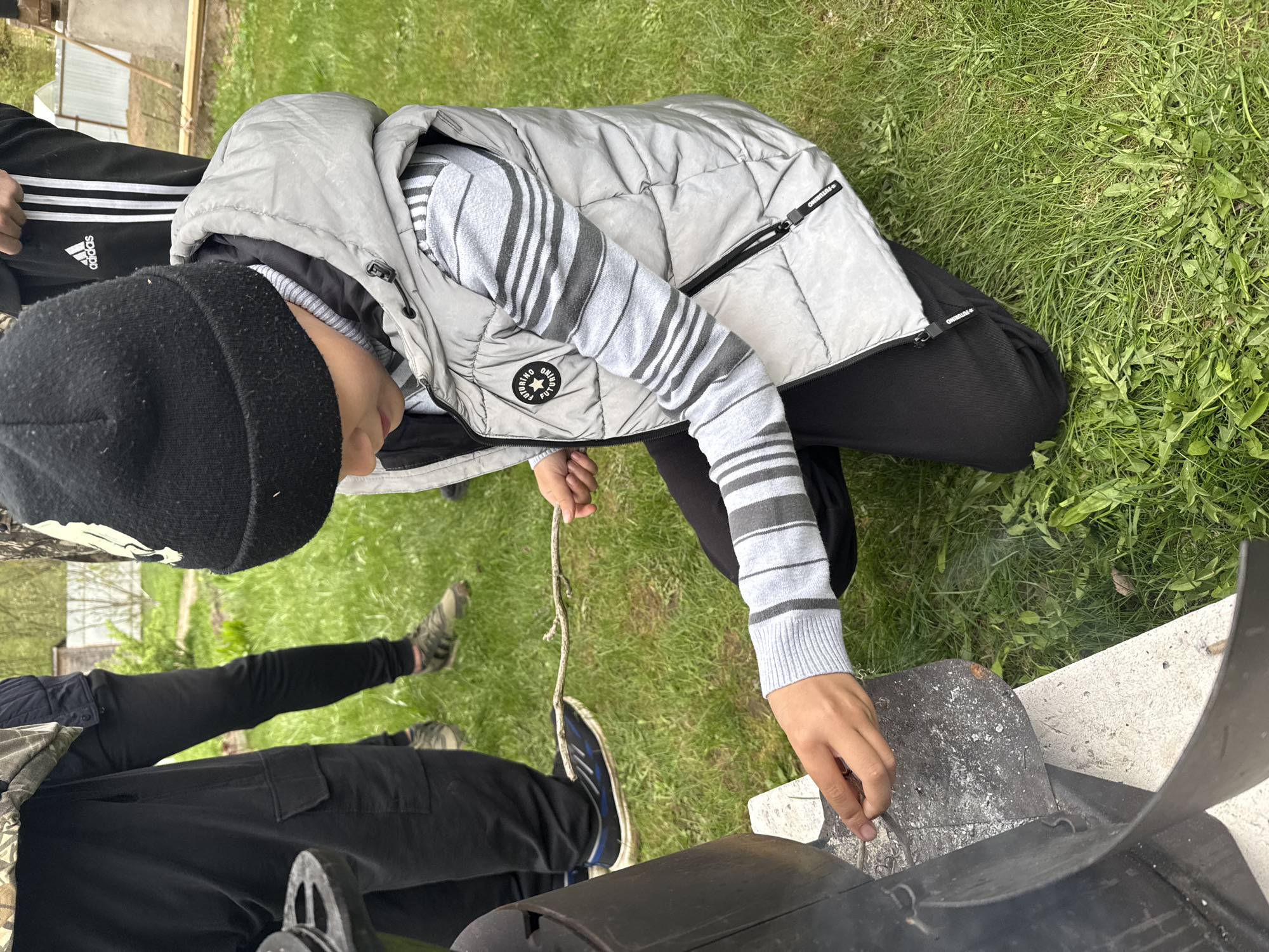 A child in a gray puffer vest, black beanie, and black pants tends to a barbecue grill in Dmitrov, Russia. Another individual in Adidas-branded pants is partially visible. The barbecue grill is black and sits on a concrete base. Green grass surrounds the area. The child is holding a stick, appearing to stir or adjust items within the grill. Smoke is visible, indicating the grill is in use.