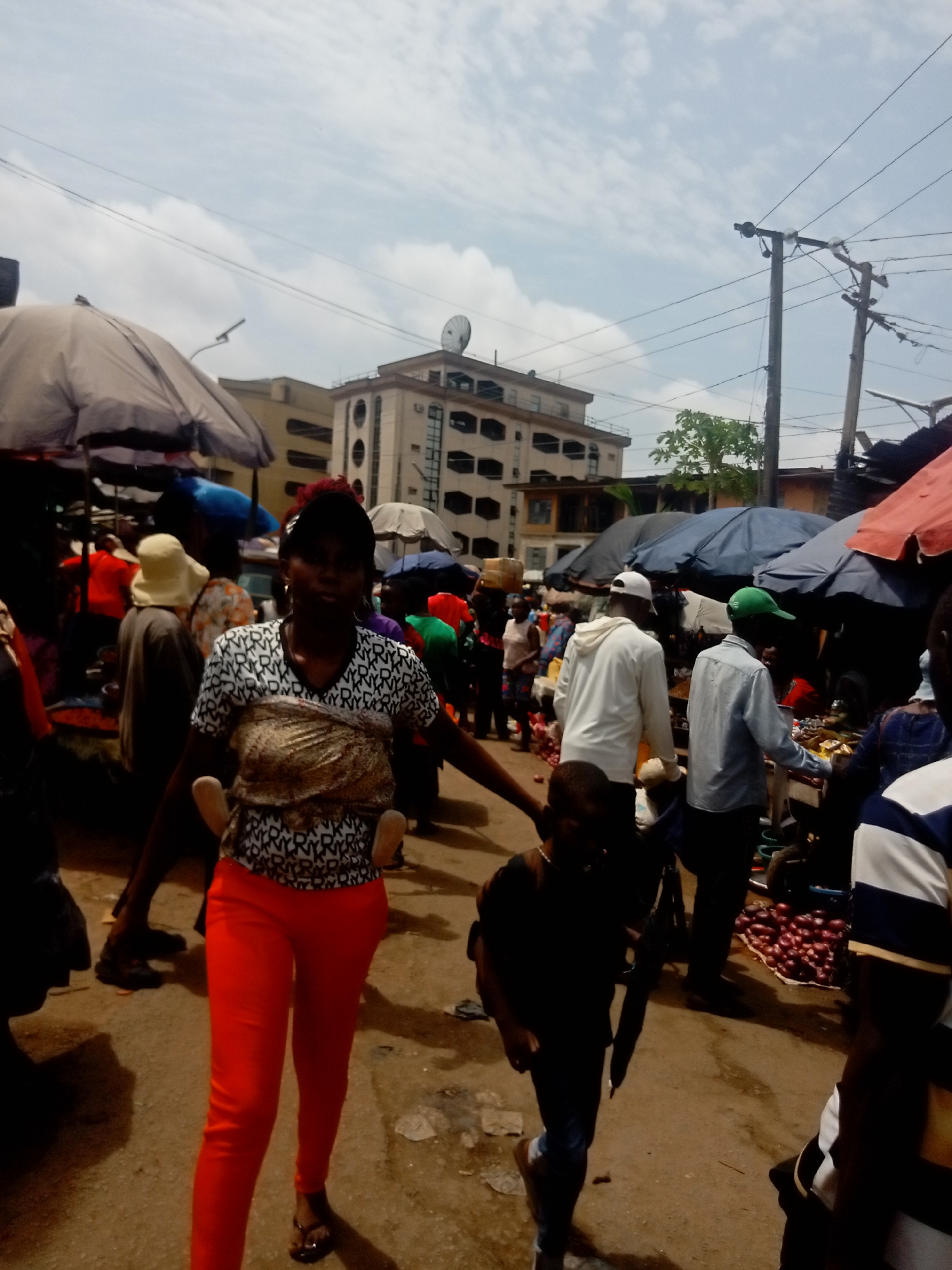 The photograph shows a bustling street scene in Onitsha, Nigeria, populated with people and vendors. A woman in a black-and-white patterned shirt and bright orange pants is carrying a child on her back in a brown cloth wrap and holding the hand of a young boy dressed in dark clothing. They are walking on a dirt and cracked pavement. Around them are other people, many walking among stalls shaded by umbrellas. Some vendors are selling goods, including red onions, on the right. Buildings are visible in the background, one with a satellite dish. Electrical wires traverse the sky above. The lighting indicates daytime.