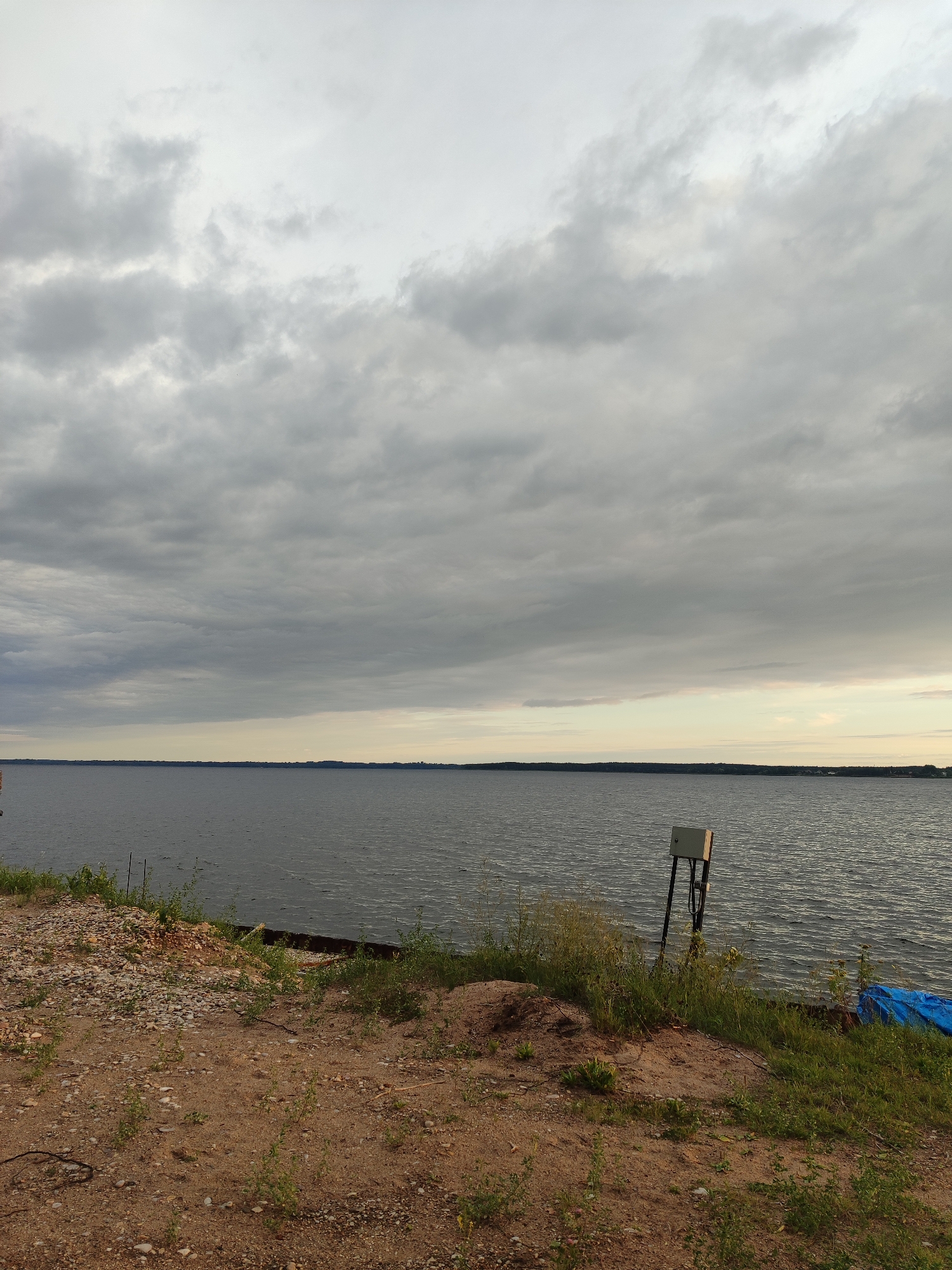 The image depicts an outdoor scene featuring a large body of water under a cloudy sky. In the foreground, there is a sandy area with scattered rocks and sparse green vegetation. A small, grey electrical box is mounted on a black pole near the water's edge. To the right, a piece of blue tarp is visible. The water stretches out to a distant horizon line, beyond which a faint outline of land with trees and buildings can be seen. The sky is filled with various shades of grey clouds, with some brighter areas suggesting diffused sunlight. The overall impression is of a natural landscape under overcast weather conditions.