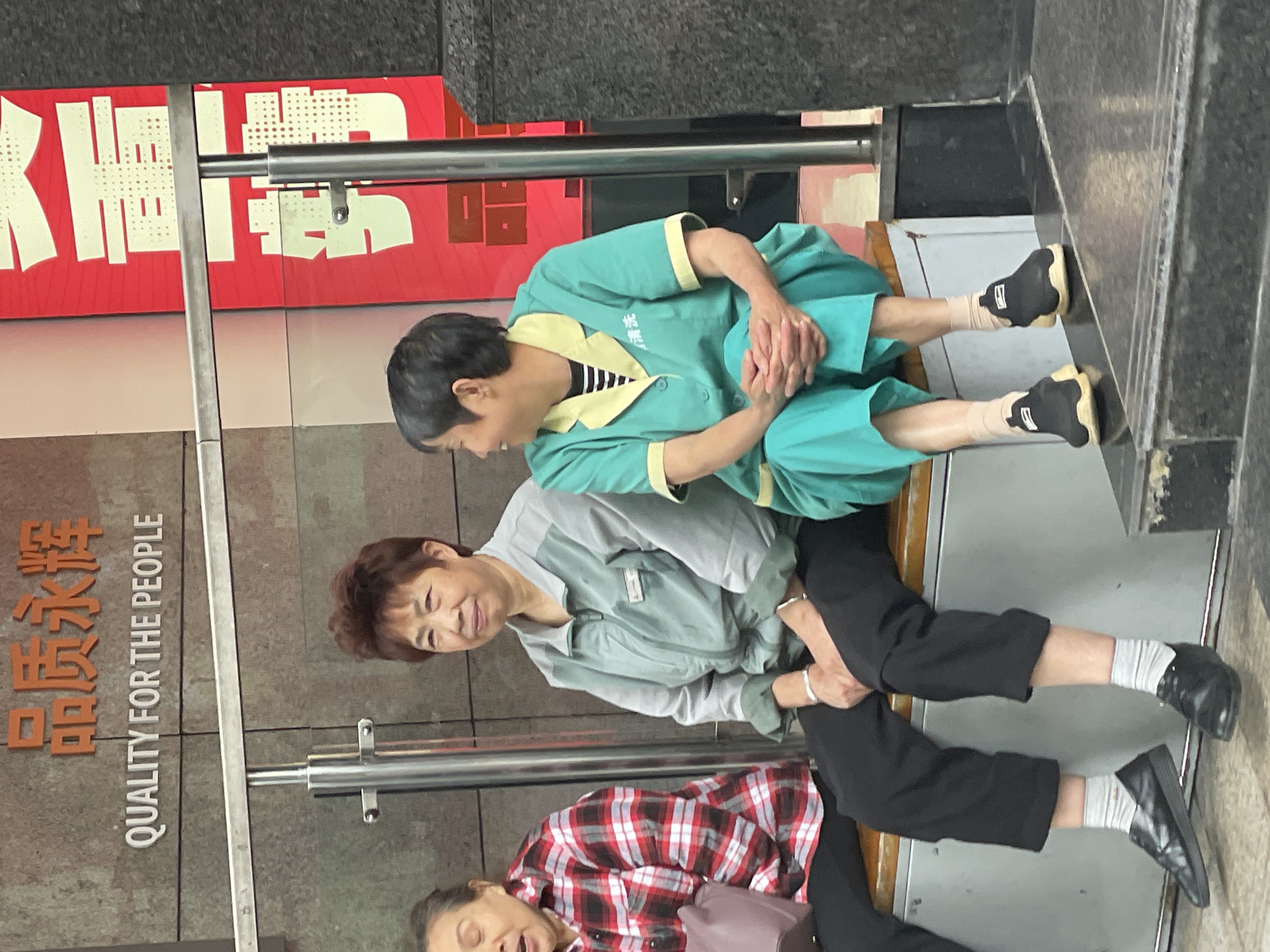 Three elderly women are sitting on a bench in Yuzhong District, China. The woman at the top is wearing a green uniform with yellow trim. The woman in the middle is wearing a light gray jacket with black pants. The woman at the bottom is wearing a red and white plaid shirt with black pants and has a purse. The bench is in front of a wall with orange Chinese writing and the English translation "QUALITY FOR THE PEOPLE." Above the women, there is a red sign with white Chinese writing.
