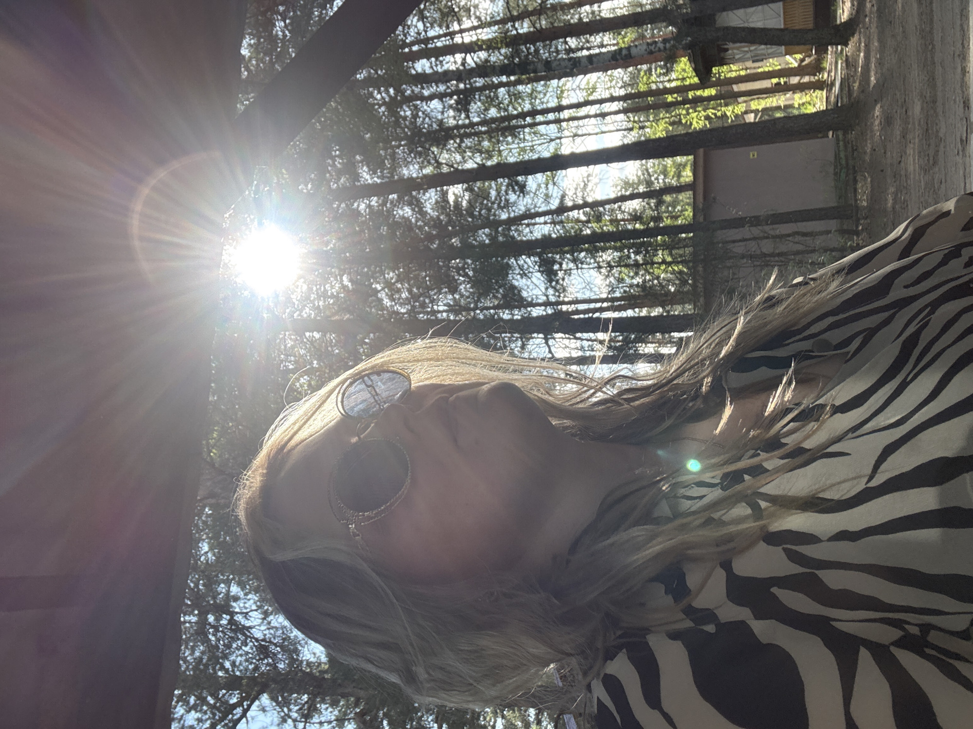 A medium-shot of a light-skinned person with blond hair and wearing dark glasses and a zebra-striped shirt. They are standing outside in a wooded area with sunlight filtering through the trees. A building is visible in the background. There is no mention of the city of Chaykovsky, Russia.