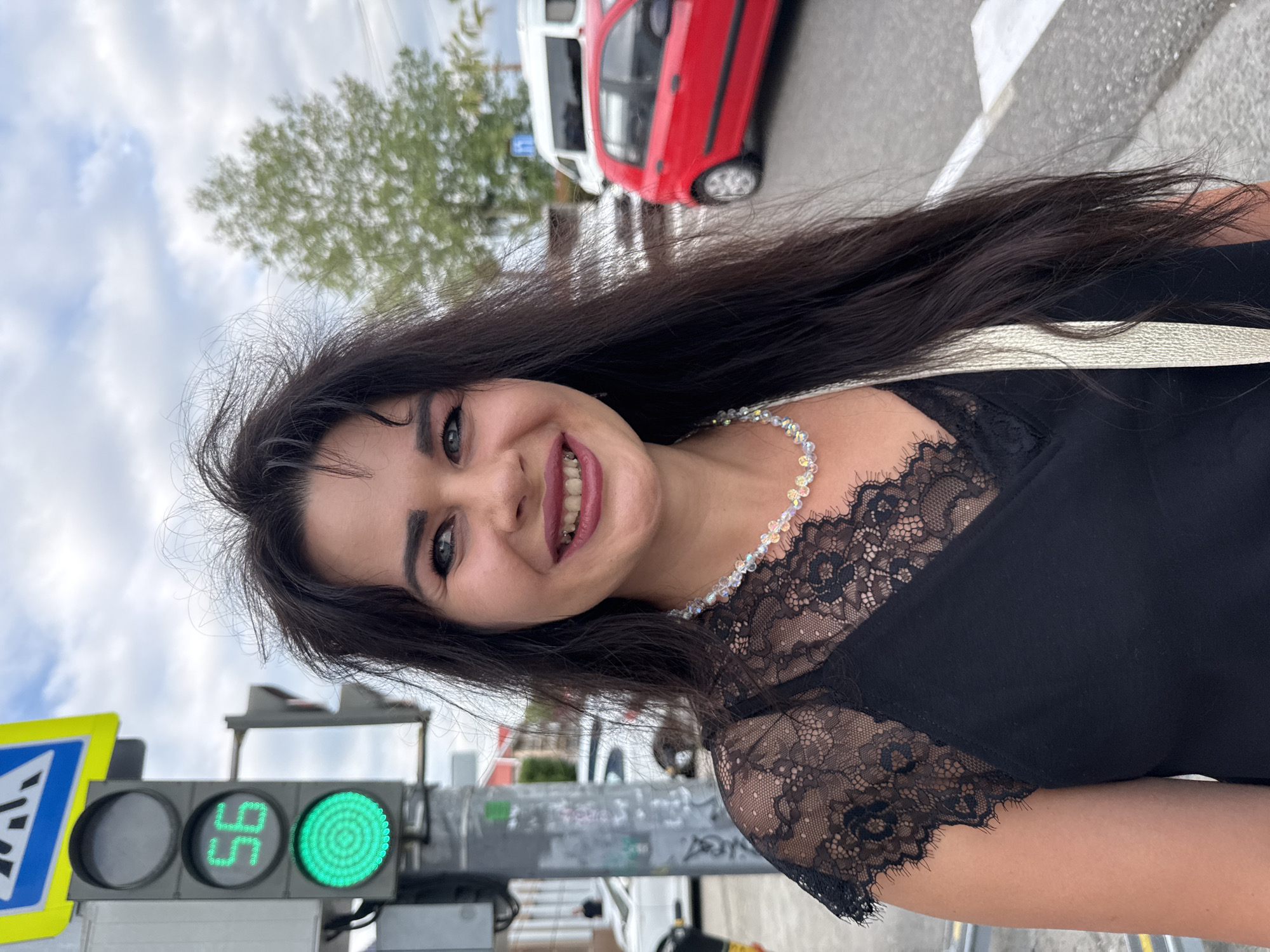 A woman with long, dark hair and blue eyes smiles into the camera. She is wearing a black top with lace detailing and a beaded necklace. The image appears to be taken outdoors in daylight, with a cloudy sky overhead. In the background, a red car and a white vehicle are visible, as well as a traffic light that is currently green. A pedestrian crossing sign is also present. The scene suggests an urban or suburban setting. The woman's expression is happy and friendly.