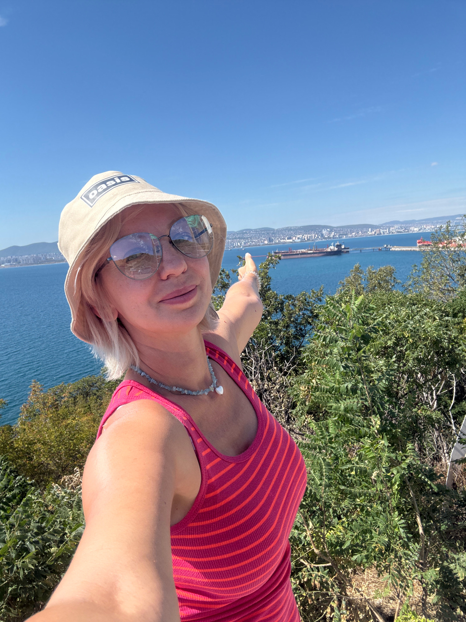 A woman wearing a pink and red striped tank top, a light-colored bucket hat with the word "Oasis" on it, and sunglasses, takes a selfie with a city skyline and ships in the background. She is standing on a hill covered in green foliage, overlooking a body of water. Her right arm is extended, pointing towards the city. The sky is a clear blue. The city appears to be Novorossiysk, Russia, characterized by its port facilities and coastal development.