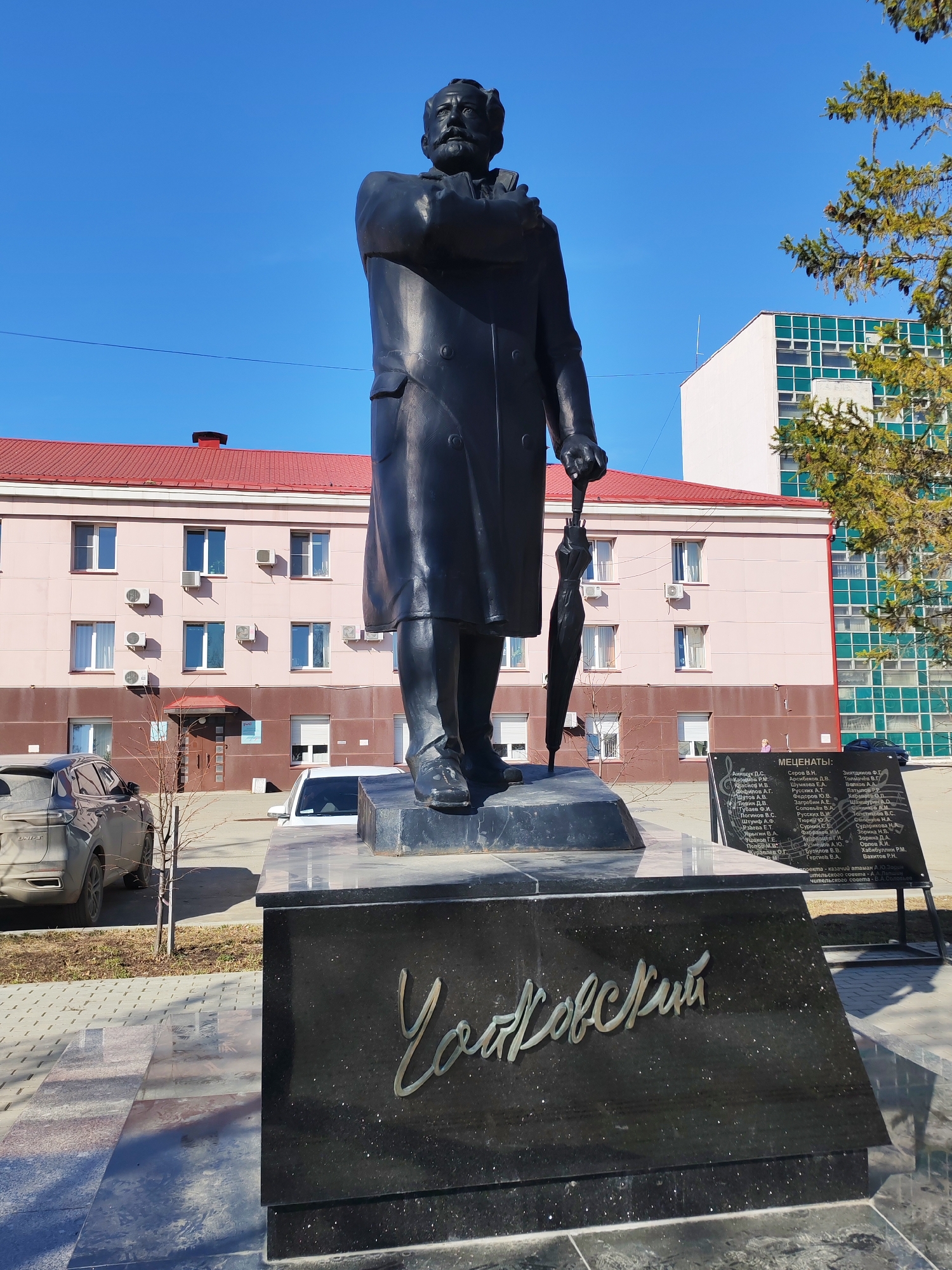 A low-angle, full shot captures a dark-colored statue of a man, likely Pyotr Ilyich Tchaikovsky, against a clear blue sky, set in what appears to be Izhevsk, Russia. The statue depicts a man with a mustache and beard, wearing a long coat, holding an umbrella in his left hand and what looks like a book or score in his right, which is resting against his chest.
The statue stands on a dark, polished stone pedestal. The front of the pedestal features the name "Чайковский" (Tchaikovsky) in stylized gold lettering.
In the background, a light pink building with a red roof and numerous windows, some with air conditioning units, is visible directly behind the statue. To the right of this building, a modern, taller building with white walls and a grid of green-framed windows can be seen. Sparse trees, some with green needles, frame the scene on the right.
To the right of the statue, a dark information board, titled "МЕЦЕНАТЫ:" (PATRONS:), lists numerous Russian names in columns, indicating contributors or sponsors. Below the names, partial text "проекта - казачий атаман А.10.Зяблов" (project - Cossack ataman A.I. Zyablov) and "ительского совета - В.А.Соловьев" (council - V.A. Solovyov) is visible.
Two cars are parked on the left side of the frame: a dark SUV partly visible in the lower left and a white car further back. The ground is paved with stone tiles and some bare earth. The bright sunlight and absence of clouds suggest a clear, possibly spring or autumn day.