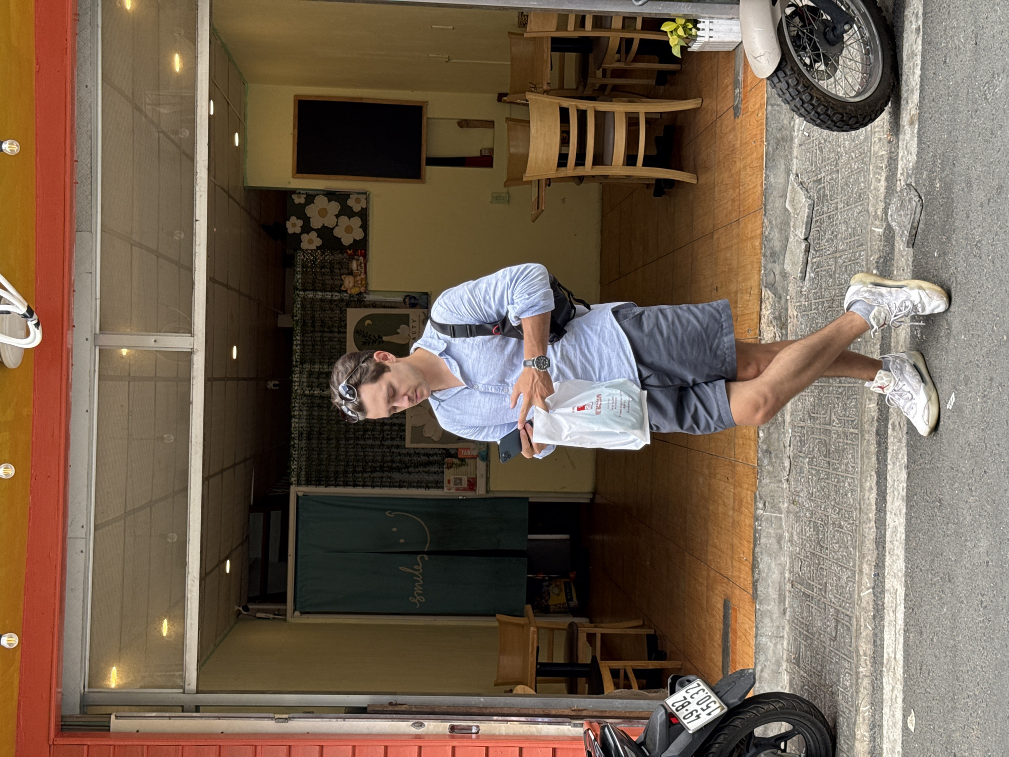 A man stands on the sidewalk in front of a business. He is light-skinned, wearing a light blue button-down shirt, gray shorts, and white sneakers. A black bag is slung across his body, and he holds a white plastic bag and a phone in his hands. Sunglasses are perched on top of his head.
The business behind him has an orange and red exterior. A screen door leads into the establishment. Inside, wooden tables and chairs are visible. Green curtains with the word "smiles" written on them in cursive block the view further into the business. A blackboard hangs on the wall.
A motorcycle is parked on the sidewalk in front of the business, partially obscuring the lower right corner of the image. The motorcycle's license plate reads "49-B2 150.32." Another motorcycle is partially visible near the top of the frame. The sidewalk appears to be textured stone, and the street is asphalt. This image was most likely taken in the city of Da Lat, Vietnam.