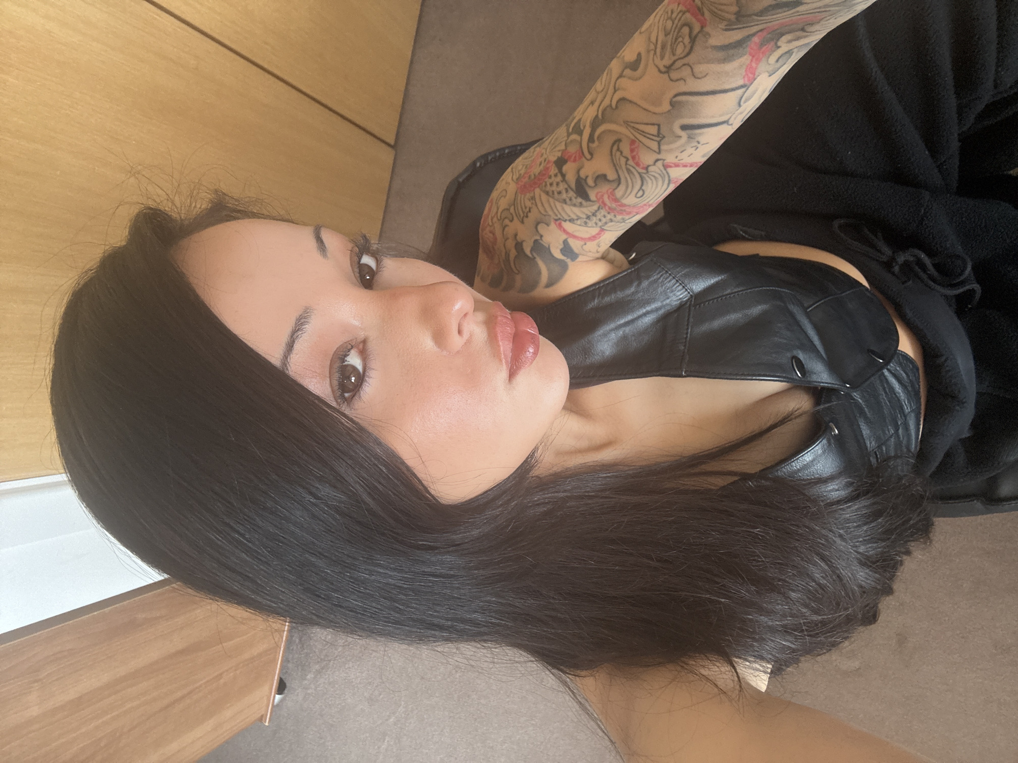 A young woman with long, straight dark hair and light brown eyes is the central subject of this close-up, angled selfie. She has fair skin and is wearing a black faux leather vest or crop top, paired with dark, possibly black, casual bottoms like sweatpants. Her left arm is heavily tattooed with a detailed Japanese-style design featuring a dragon or koi fish amidst waves and clouds, rendered in black, grey, and red.
She is posing with her head tilted, and her lips are slightly pursed in a "duck face" expression, looking directly at the camera. Her eyebrows are well-defined, and she has visible mascara or false eyelashes, along with a glossy, natural-toned lip color. A few wispy hairs are visible around her hairline.
The setting appears to be indoors. To her left, there's a light wooden surface, possibly a wall or furniture. To her right and below, there's a dark grey or brown floor or carpet. The lighting suggests it is daytime, with a soft, natural light source illuminating her face. No specific London cues are visible within the image, though the context indicates the location is London, United Kingdom. There is no visible text in the image.