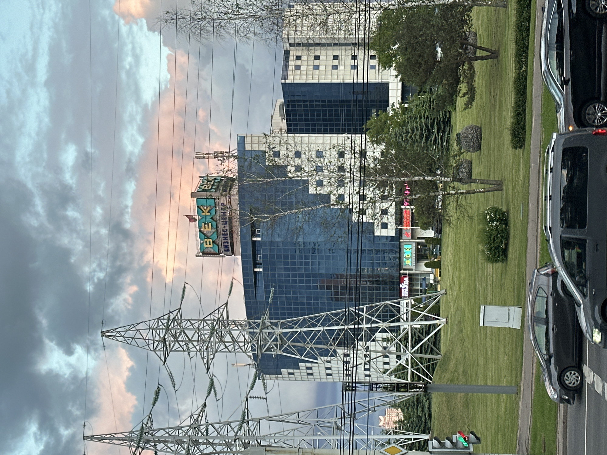 The image shows a view of a city street, likely in Minsk, Belarus, given the Cyrillic text "ВЕК Бизнес-центр" (Vek Business Center) on a sign atop a building. In the foreground, there is an electrical transmission tower, which is partially obscuring the view of the building. The modern building behind the tower has a grid-like pattern on its blue glass facade. To the right of the building, there is a green area with trees and shrubbery. Cars are parked on the side of the road, indicating a busy urban environment. A traffic light is present. The sky above is cloudy.