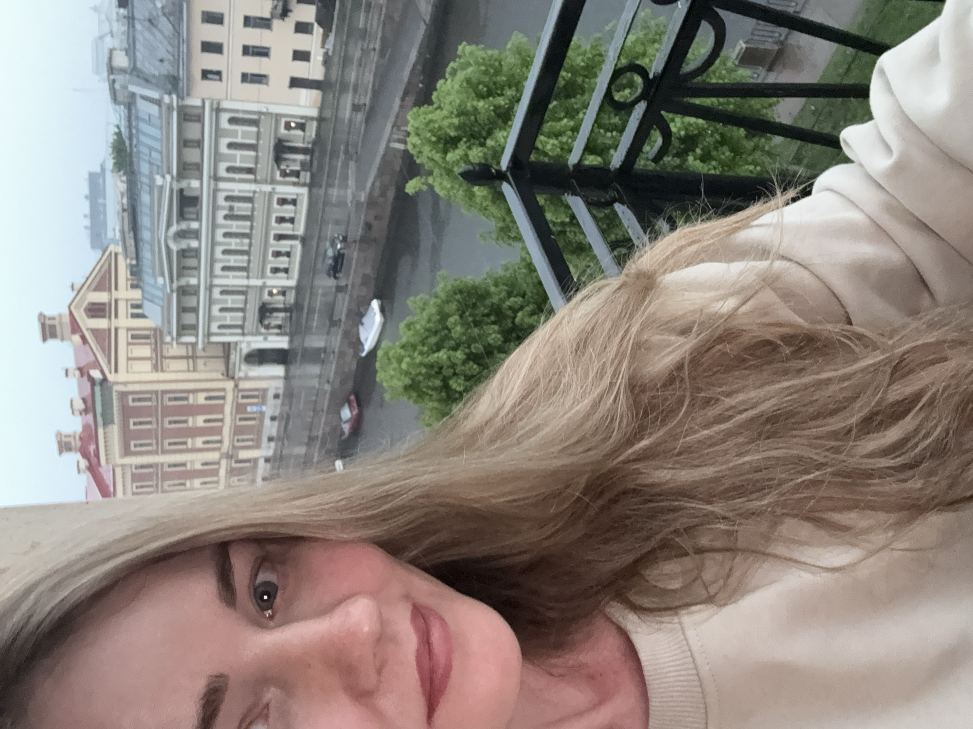 A woman is taking a selfie on a balcony overlooking a river in Saint Petersburg, Russia. The buildings across the river are visible. The weather appears overcast and possibly rainy. A black iron railing is in the foreground. The woman has blond hair and is wearing a light-colored sweatshirt.