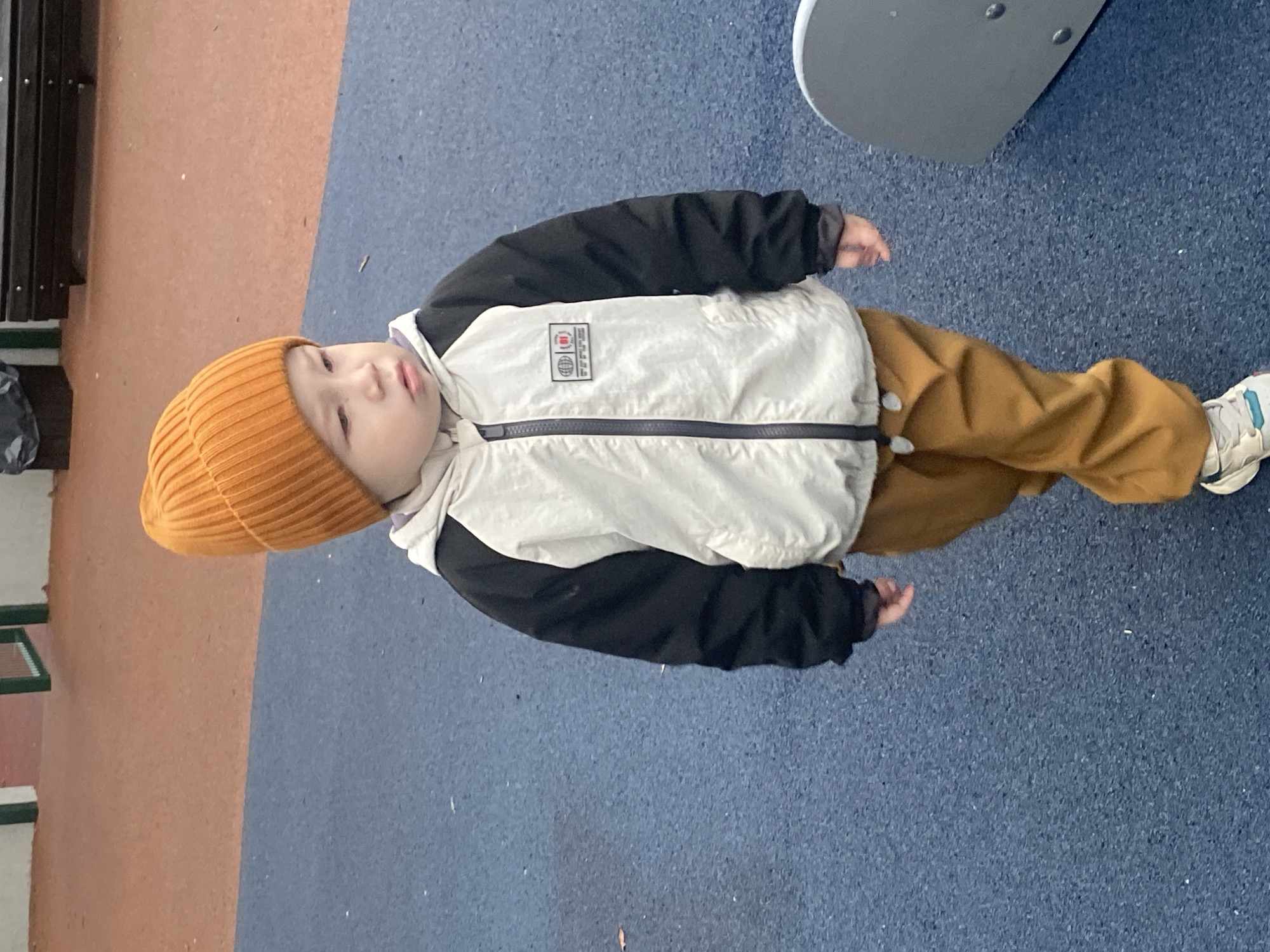 An outdoor, full-body shot captures a young child standing in what appears to be a playground or park in Sochi, Russia. The image is rotated counter-clockwise, showing the child standing upright on a blue, speckled rubberized safety surface, with a section of an orange-brown textured ground visible to the left.
The child, likely a toddler or preschooler, is dressed in warm winter or cool-weather clothing. They wear a bright orange knitted beanie, a two-tone jacket with a light cream or white body and black sleeves, brown pants, and white sneakers with hints of blue or green. Their left hand is visible at their side. The child's gaze is directed slightly to the left of the frame, with a neutral, slightly pensive expression and lips slightly parted.
In the background, a dark structure, possibly a bench or fence, is partially visible on the far left. On the far right, a light grey, curved metal object, likely part of playground equipment, is present.
On the upper left chest of the child's jacket, a rectangular patch is visible. It contains various graphic elements including what appears to be a stylized globe icon, possibly the word "LIFE" stacked vertically, and other small, indistinct symbols or text. The overall scene suggests a cool, daytime outing.