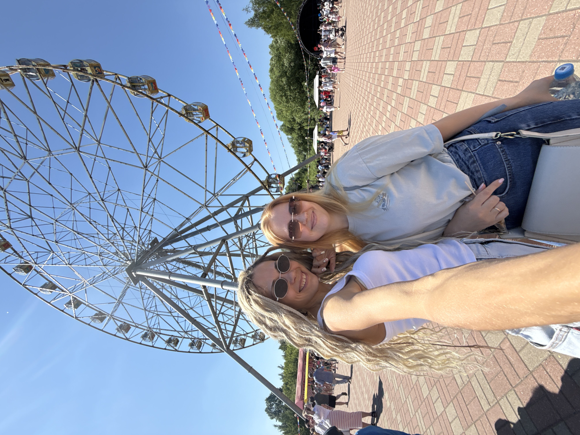 Two young women are taking a selfie in an outdoor amusement park. The woman on the left has long blonde dreadlocks, wears a white tank top and sunglasses. The woman on the right has long red hair and wears a light blue t-shirt and sunglasses. Behind them, a Ferris wheel is visible against a clear blue sky. In the background, there are trees, a stage with a black canopy, and a crowd of people walking around. The ground is paved with red and beige bricks. The scene appears to be the amusement park in Zavolzhye, Russia, on a sunny day.