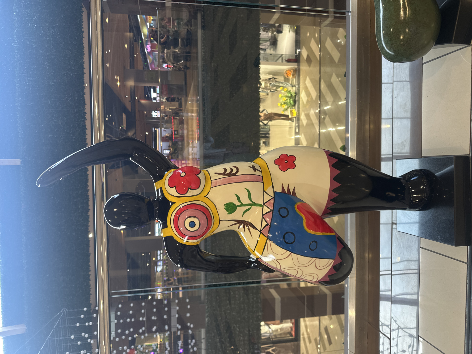 A glossy black sculpture of a stylized female figure, adorned with bright, colorful, abstract patterns, stands indoors, possibly in a shopping mall. The figure is positioned facing forward, with its left arm raised and its right arm bent at the elbow and held close to its body. The torso is painted with circles resembling targets and a floral motif, while the lower body features geometric patterns and a blue and red shape. The sculpture is placed on a black rectangular base, which rests on a tiled floor. Behind the sculpture, a glass partition reveals a multi-level interior space with shops, escalators, and people in the background, suggesting a commercial environment. The overall lighting is bright, with light reflecting off the sculpture's surface and the polished floors. The location provided in the prompt, Stavropol, Russia, is not ascertainable from the image itself.
