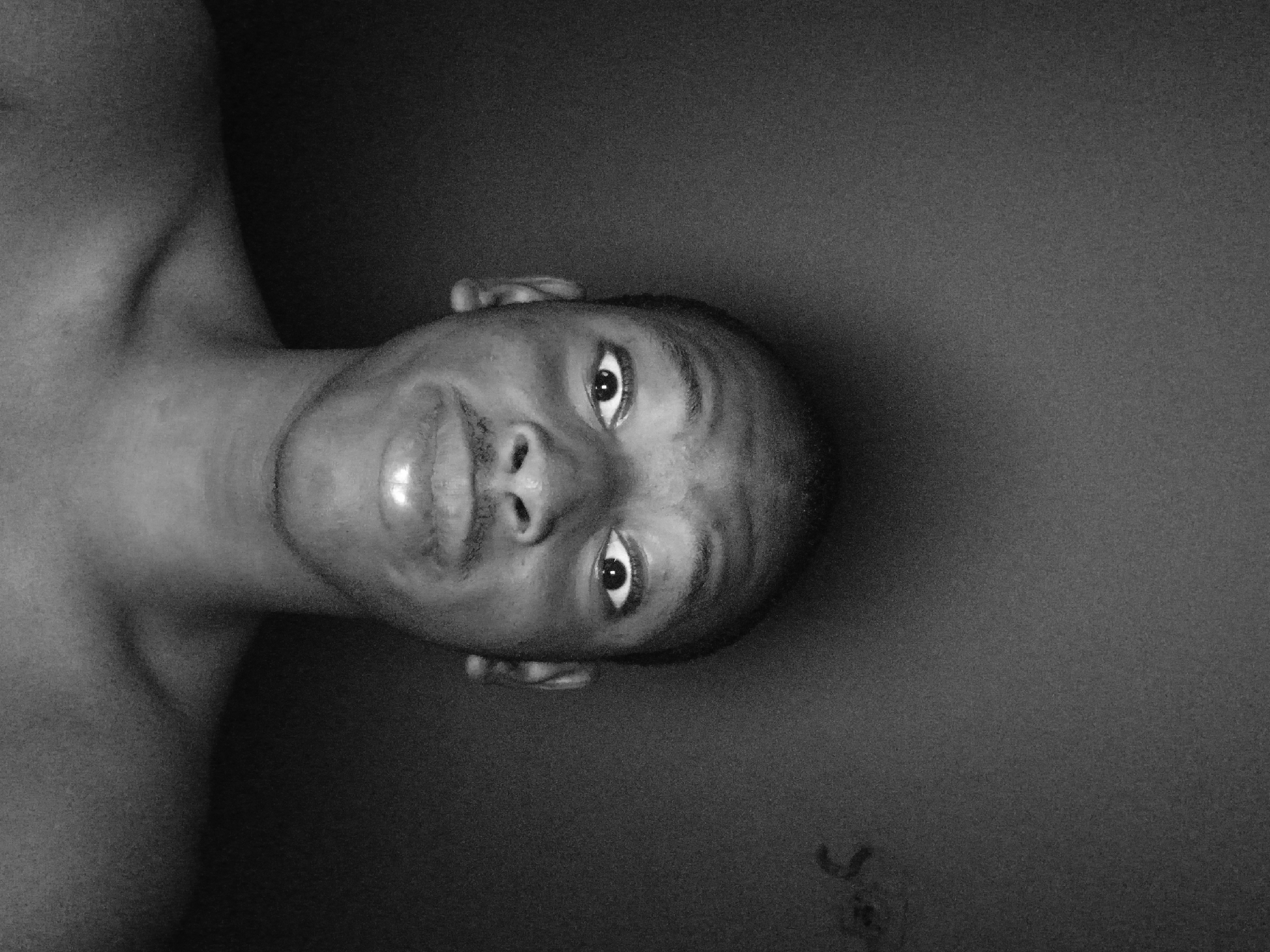 A close-up, black and white photograph depicts a man’s head and partial shoulder. He has dark skin, a shaved head, and is looking directly at the camera. His facial expression appears neutral. Part of his bare shoulder is visible in the left of the image. The background is a dark, uniform tone. The image appears to be taken in Lagos, Nigeria. There is nothing to indicate the location.