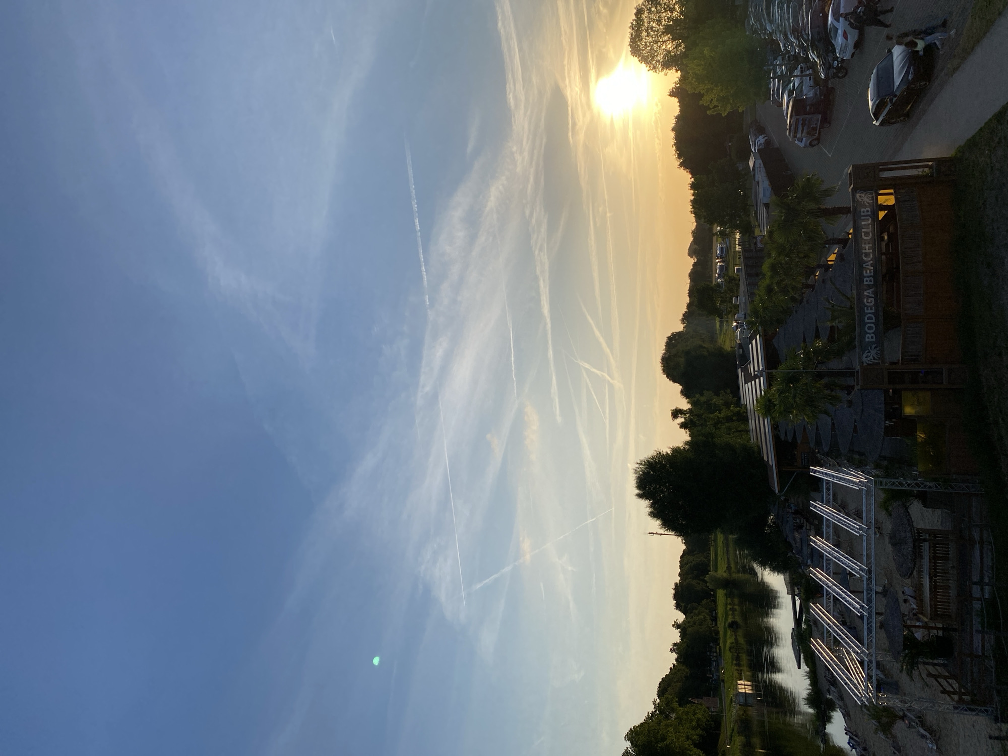 The image depicts a view of the Weser riverfront in Rinteln, Germany, at sunset. In the foreground, a beach club with thatched umbrellas and seating areas is visible. A sign reads "BODEGA BEACH CLUB". Adjacent to the beach club is a parking lot filled with numerous cars. The Weser river flows to the left of the beach club, with trees lining its banks. In the sky, a bright sunset casts a warm glow, with scattered clouds and contrails from aircraft. A small, bright green circular object is visible in the upper left quadrant of the sky. Several people are present in the scene, walking near the parked cars and on the beach.