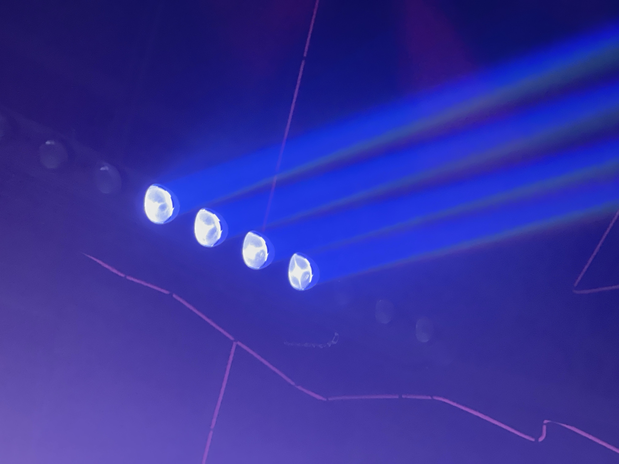 The image depicts a row of spotlights emitting bright blue beams into a dark, purple-toned space. There are at least four discernible spotlights on the left side of the image. The beams of light are slightly separated, showing horizontal striations within the blue light. There is an object on the ground. There is some pink string lighting.
The image location is not discernible.