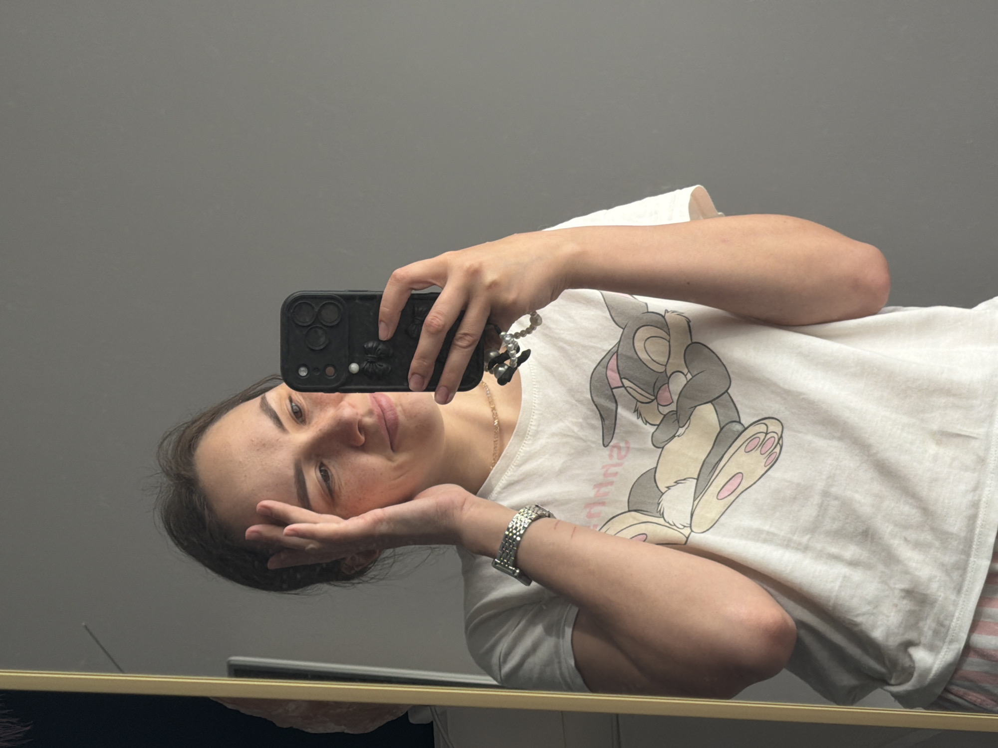 A young woman is taking a selfie in a mirror. She is wearing a white t-shirt with a cartoon bunny design and a silver watch. She holds a black smartphone with a decorative charm attached. The background appears to be a plain, grey wall. The location is assumed to be in Chaikovsky, Russia.