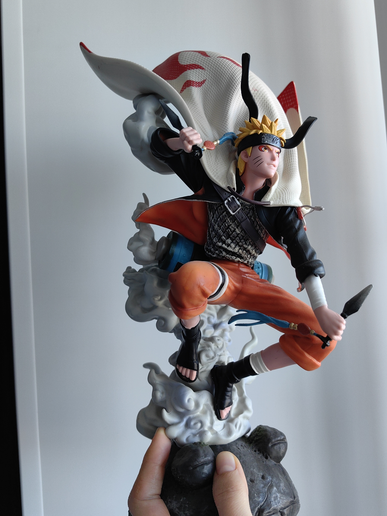 A person holds a Naruto Uzumaki figurine in their hand. The figurine depicts the character in a dynamic pose, wielding a kunai and leaping from a cloud-like base. He wears an orange outfit, a black top, and a white cape. The setting appears to be indoors, with a plain white background. No city, country, or other location details are evident in the image.