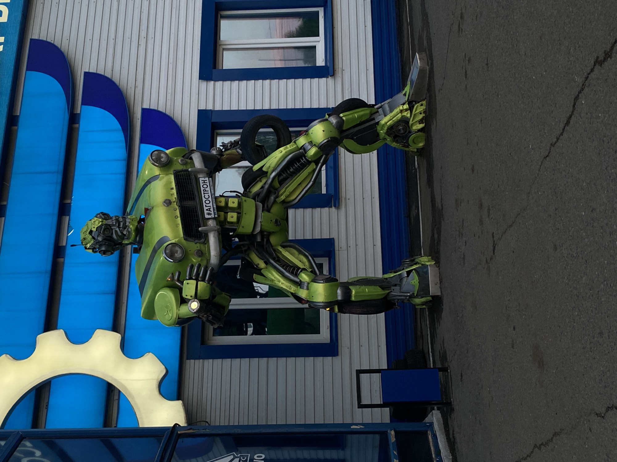 An outdoor shot captures a large, vibrant green robot sculpture standing in front of a white building with blue accents. The robot is intricately designed, resembling a Transformer, with visible mechanical parts and a car hood forming its torso. A tire is mounted on its right arm, and its left hand holds a small object that appears to be emitting light. A license plate with the text "#АГОСТРОН" is affixed to the robot's chest. The building behind the robot features horizontal siding and several windows. To the left of the robot, a large, golden gear-shaped cutout is partially visible. The ground in front of the robot is paved with asphalt, showing some cracks. The scene is set in the city of Khokhryaki, Russia. There are no people visible in the image.
