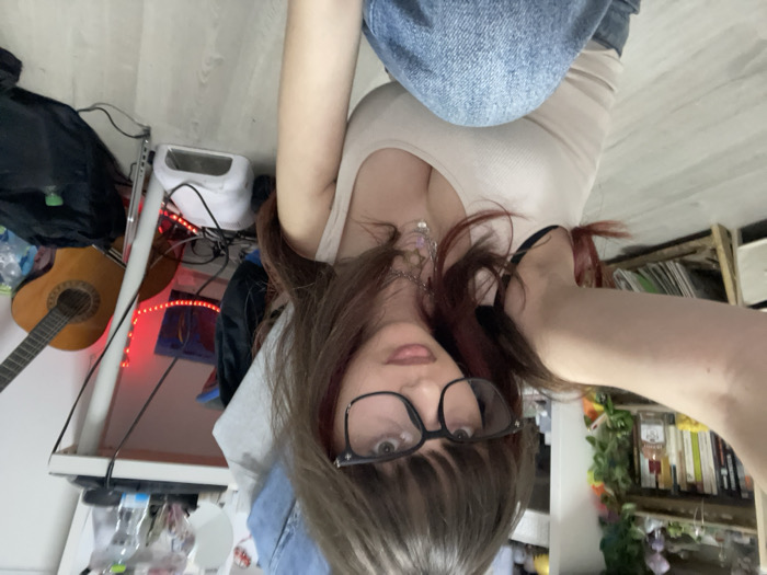 A person with glasses, a beige tank top, and denim clothing is the focus. The background is cluttered. A guitar leans against a white table with a toaster, water bottles, and a black bag. LED lights are visible. A bookshelf with books, plants, and other items stands to the right. The image is rotated, giving an upside-down perspective.