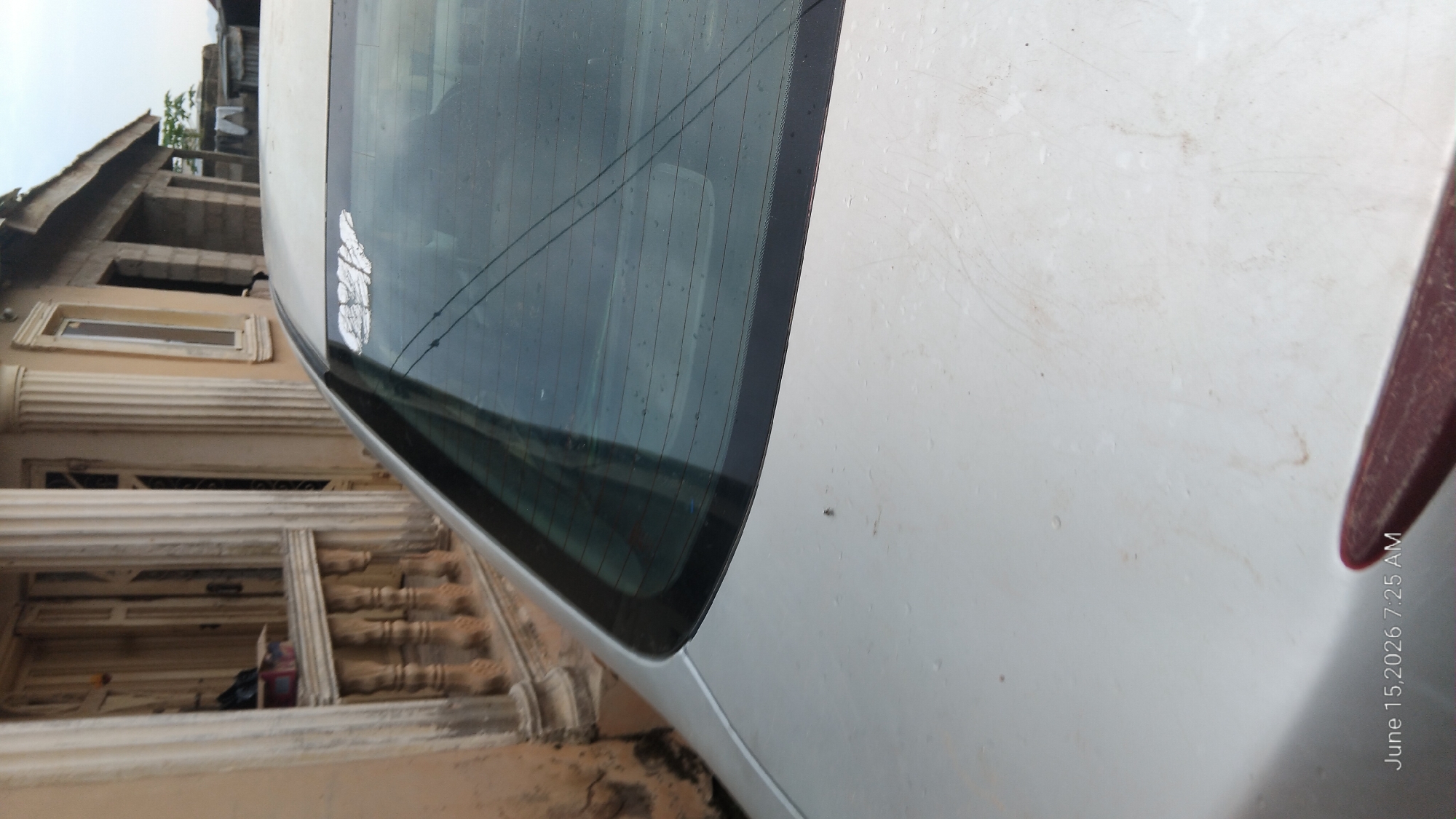 The image shows the rear of a silver car parked outdoors, with a building and a street in the background. The car has a faded sticker on the rear window. The building appears to be made of concrete and has a traditional Nigerian architectural style. There is some debris on the street and sidewalk. The time stamp on the image indicates it was taken on June 15, 2026, at 7:25 AM. The location is Howayi, Nigeria.