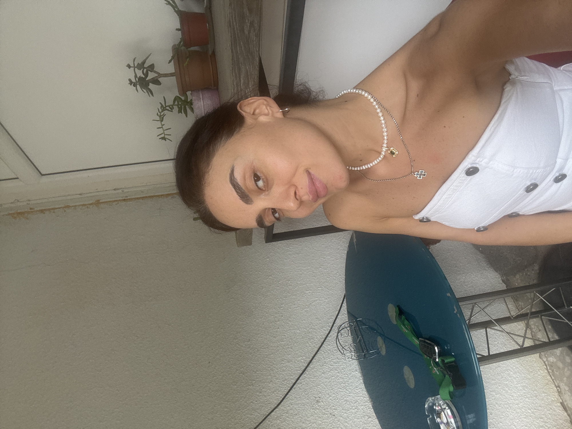 A young woman with dark hair and light skin is taking a selfie outdoors. She is wearing a white strapless top and two necklaces, one with pearls and a pendant, and another with a cross pendant. She is leaning against a wall, and a teal table is visible in the foreground with an ashtray, keys, and a lanyard on it. In the background, several potted plants are arranged on a shelf. The lighting suggests it is daytime. The architectural details of the wall and the presence of potted plants on a shelf are common features found on balconies or patios in Tbilisi, Georgia.