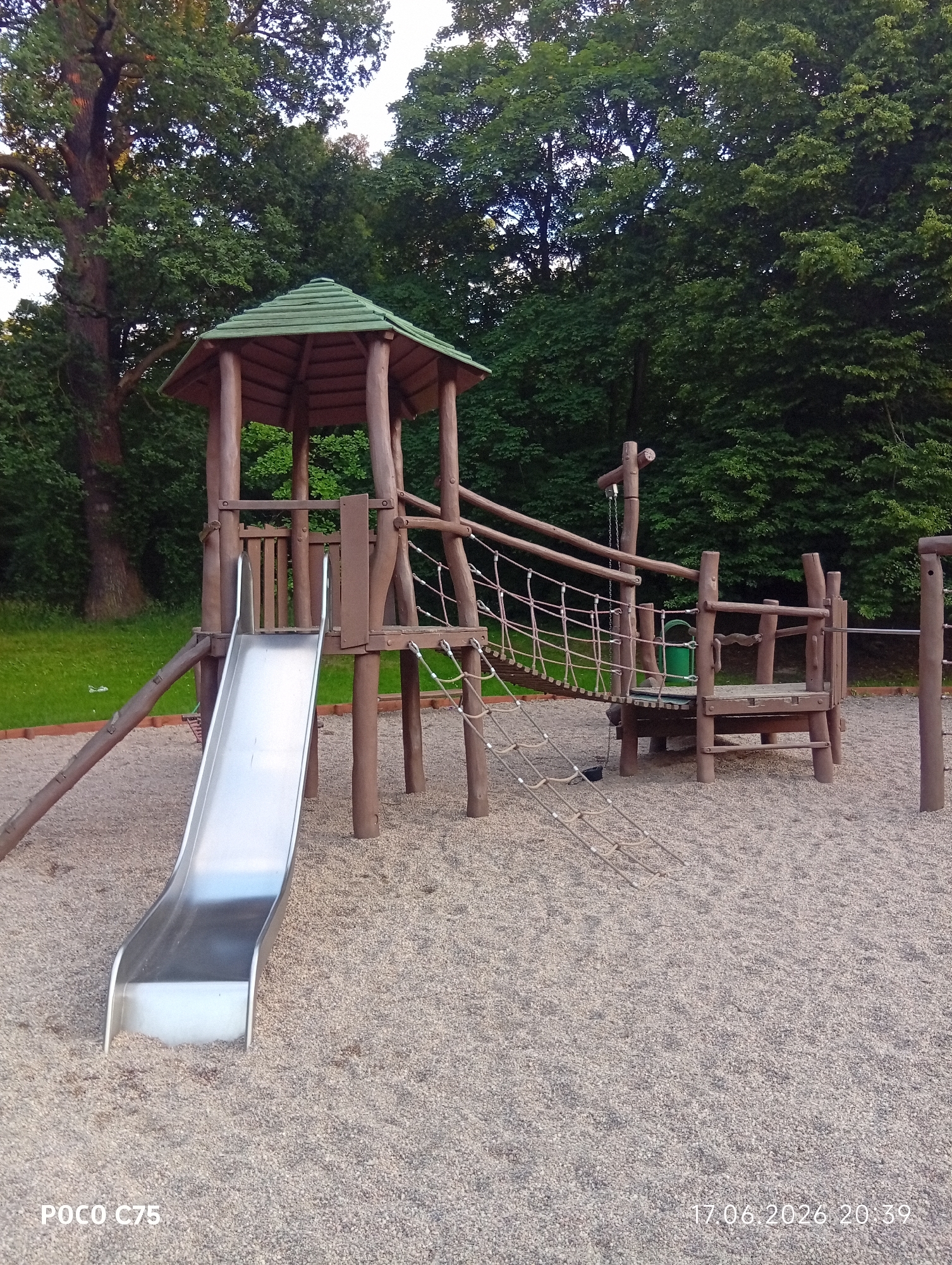 The image depicts a playground structure made of wood and metal. The main subject is a multi-level wooden play tower with a green, angled roof. A shiny silver slide descends from the tower to the ground, which is covered in small, light-colored gravel. The tower has various wooden posts, beams, and railings. A rope bridge connects the tower to another wooden platform. There are also sections of rope netting, suggesting climbing or balancing elements. A green bucket is visible hanging from one of the wooden structures.
The setting is outdoors, with lush green trees forming a dense backdrop behind the playground. The ground in front of the playground is grass, indicating a park or recreational area. The lighting suggests it is daytime, possibly late afternoon or early evening, with the sun casting soft shadows. The weather appears to be clear. The image was taken in Wrocław, Poland.
No people are visible in the image, so no activity or event involving humans is taking place. The playground appears to be empty. Notable details include the sturdy construction of the play equipment, the naturalistic setting with mature trees, and the clean, well-maintained appearance of the playground.
Visible text in the image includes "POCO C75" in the bottom left corner and "17.06.2026 20:39" in the bottom right corner, likely indicating the device used to take the photo and the date and time.