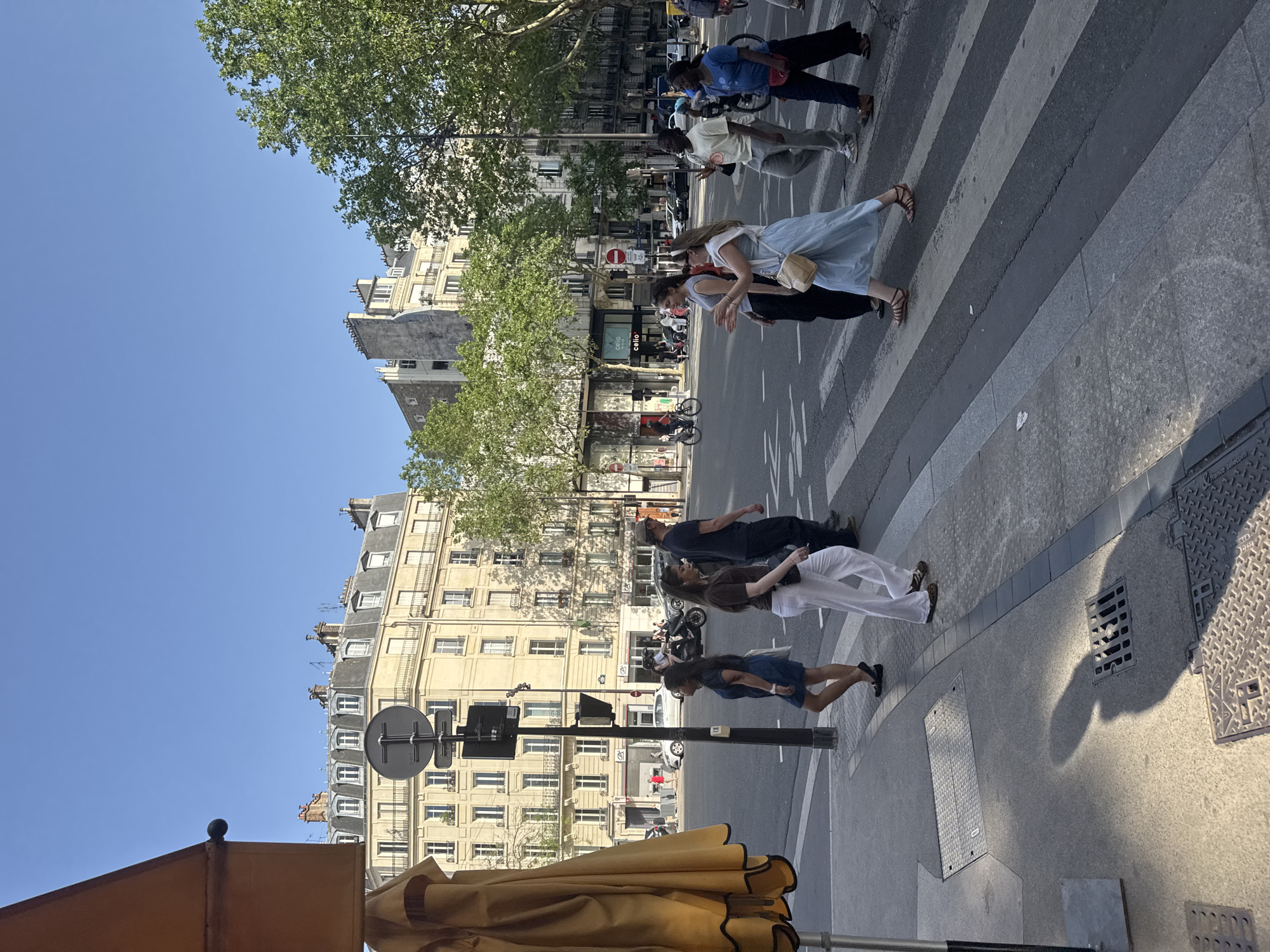 The image captures a street scene in Paris, France. The sky is clear and blue. Pedestrians are crossing a zebra-striped crosswalk on a street lined with buildings. The buildings have multiple windows and architectural details. Trees line the street, providing shade. A brown umbrella is visible in the foreground. People are walking in both directions across the crosswalk. The street appears to be paved, and there are utility covers visible on the sidewalk. A street sign is present. There are parked vehicles and motorcycles visible further down the street.