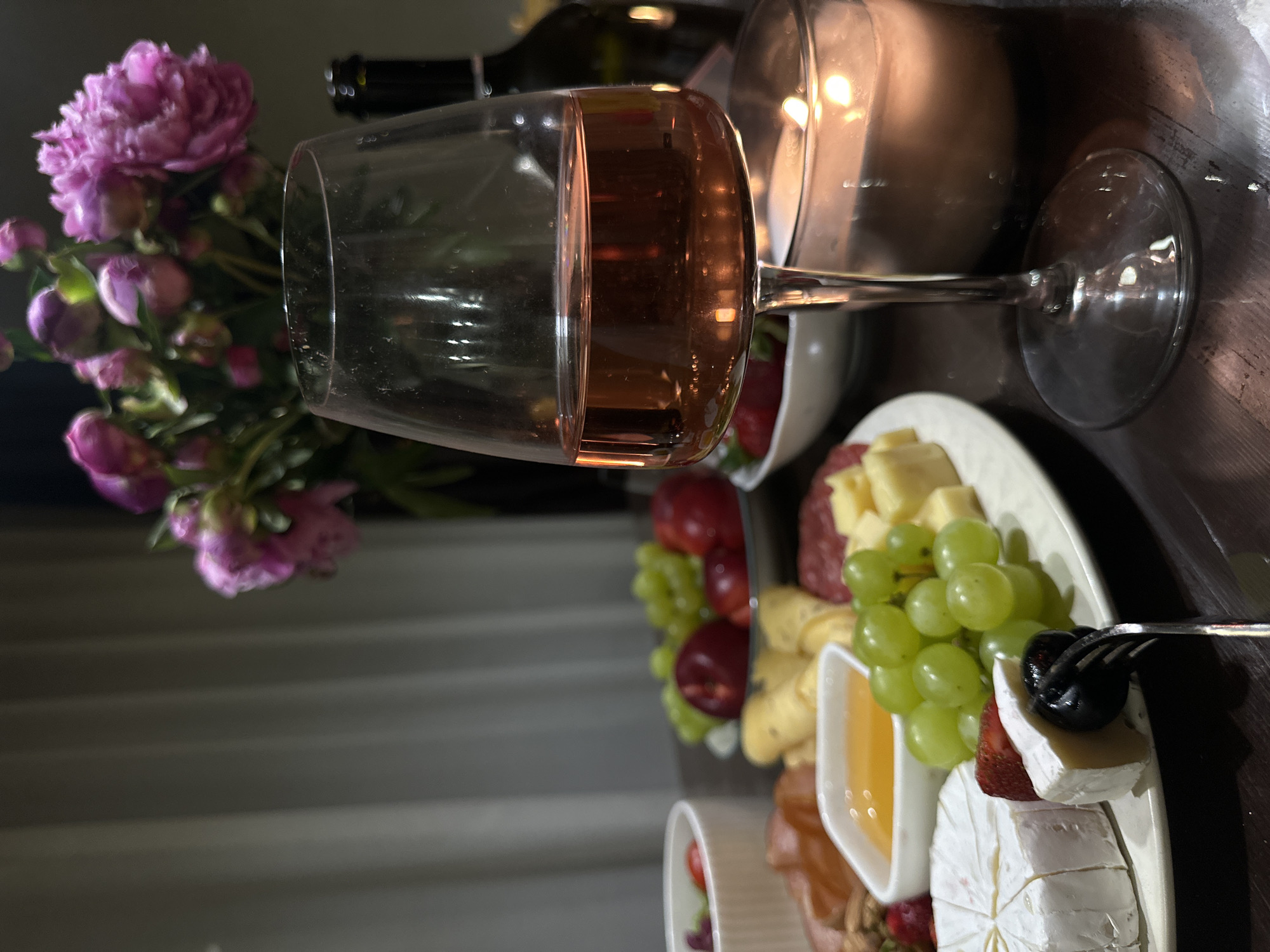 The image depicts a close-up view of a charcuterie board and a glass of rosé wine, set on a dark wooden surface. The charcuterie board is laden with various items, including wedges of brie cheese, a cluster of green grapes, slices of salami, and what appears to be honey in a small white dish. Several strawberries are also visible, some placed on the board and others in a white bowl in the background. A small fork with a dark olive is positioned on the charcuterie board. To the right of the wine glass, a lit candle in a glass holder casts a warm glow on the scene. In the background, a bouquet of vibrant pink flowers, possibly peonies, is arranged in a vase, and a dark bottle, likely of wine, is partially visible. The setting appears to be indoors, with a softly blurred curtain in the background. There are no people visible in the image. The image does not contain any information that would allow for the identification of its location in Chaikovsky, Russia.