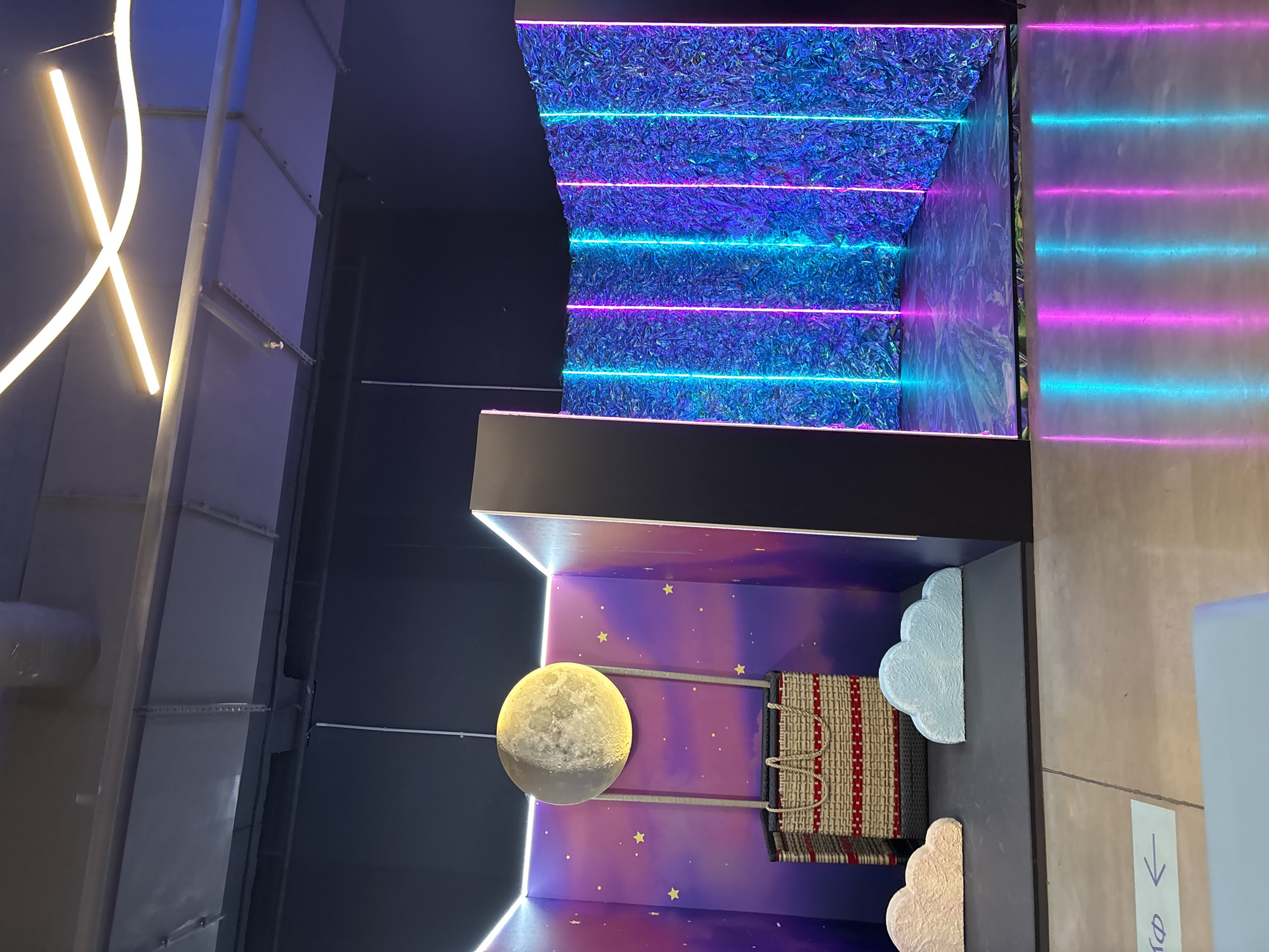 The image depicts an indoor scene with a surreal, dreamlike aesthetic. In the left section, a large, illuminated moon hangs above a wicker basket, suspended by ropes. The backdrop is a gradient of purple and pink, adorned with small yellow stars, resembling a night sky. Two stylized clouds, one pale pink and the other light blue, rest on the floor in front of the basket.
The right section of the scene features a wall covered in a shimmering, textured material with a blue and purple iridescent sheen. Vertical neon lights, alternating between cyan and magenta, illuminate this wall, creating a vibrant, futuristic atmosphere. The floor in this area has a reflective, marbled pattern that mirrors the neon lights.
The overall environment appears to be an exhibition space or a photo booth, characterized by dramatic lighting and thematic decor. The ceiling above is industrial, with visible ductwork and metallic beams, contrasting with the fantastical elements below. The floor in the foreground is a light gray concrete. A light blue sign with a black arrow pointing left and indecipherable text is visible on the concrete floor. No people are present in the image. The location is not Saint Petersburg, Russia.
