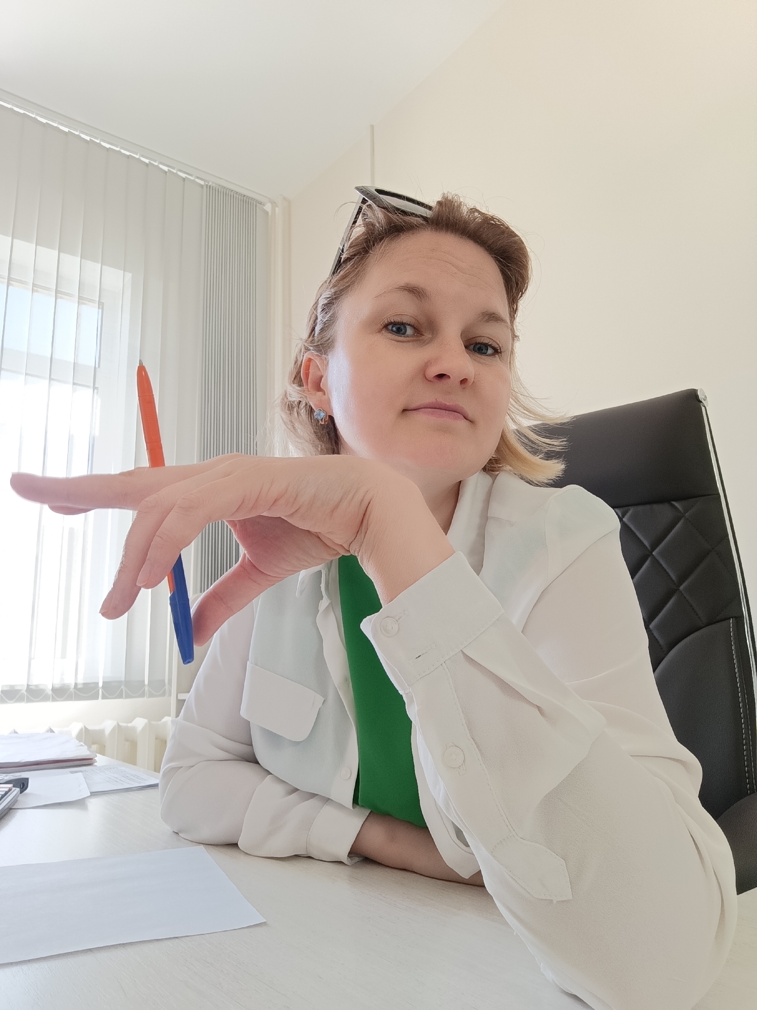 A woman with blonde hair pulled back is seated at a desk, holding two pens, one orange and one blue. She is wearing a white button-down shirt over a green garment. Her left hand, holding the pens, is raised and positioned near her face. She is looking towards the camera. The background shows a window with vertical blinds, a radiator, and a black office chair. Papers are visible on the desk in front of her. The scene appears to be an office setting.
