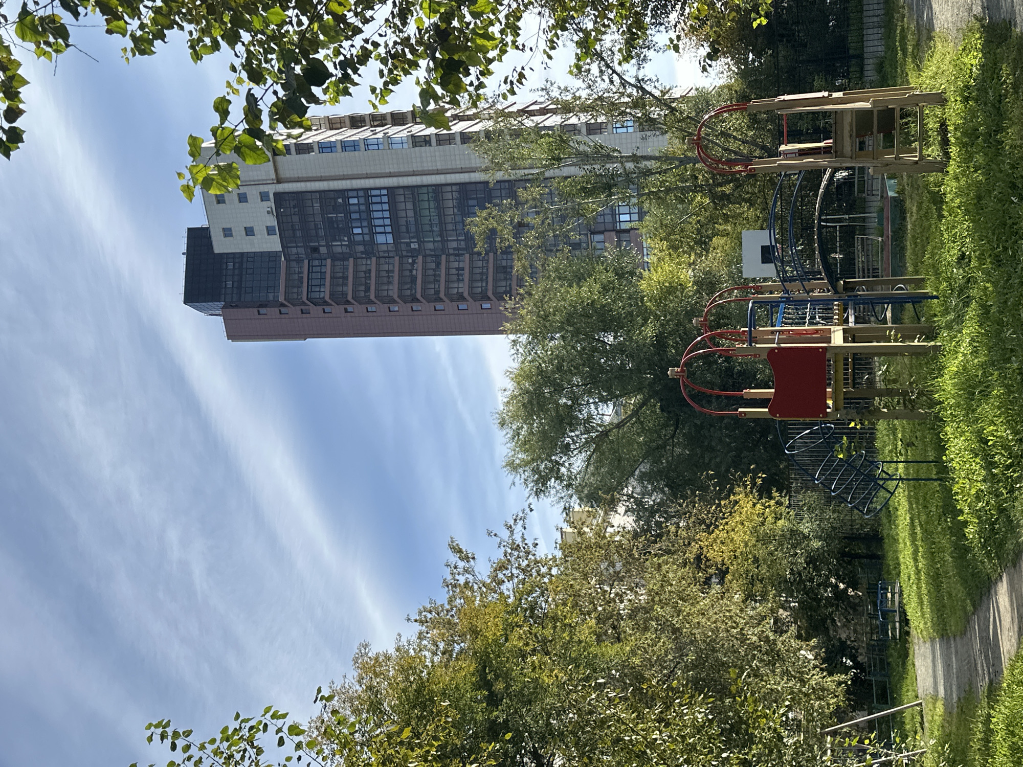 In the city of Unknown, Unknown, an outdoor scene depicts a residential complex with a towering, modern apartment building. The building features a facade with varied materials, including a dark upper section, a light-colored middle section with multiple windows, and a pinkish-brown lower section with balconies. Adjacent to the building, a playground with various equipment is visible. This includes a red and yellow play structure with a ladder, a blue spiral slide, and a small merry-go-round. The playground is surrounded by lush green grass and mature trees with foliage indicating an autumn season, with some leaves showing yellow and green hues. A paved pathway leads through the grassy area, and a black metal fence is present in the background. The sky is partly cloudy, with wispy white clouds against a clear blue background. No people are present in the image.