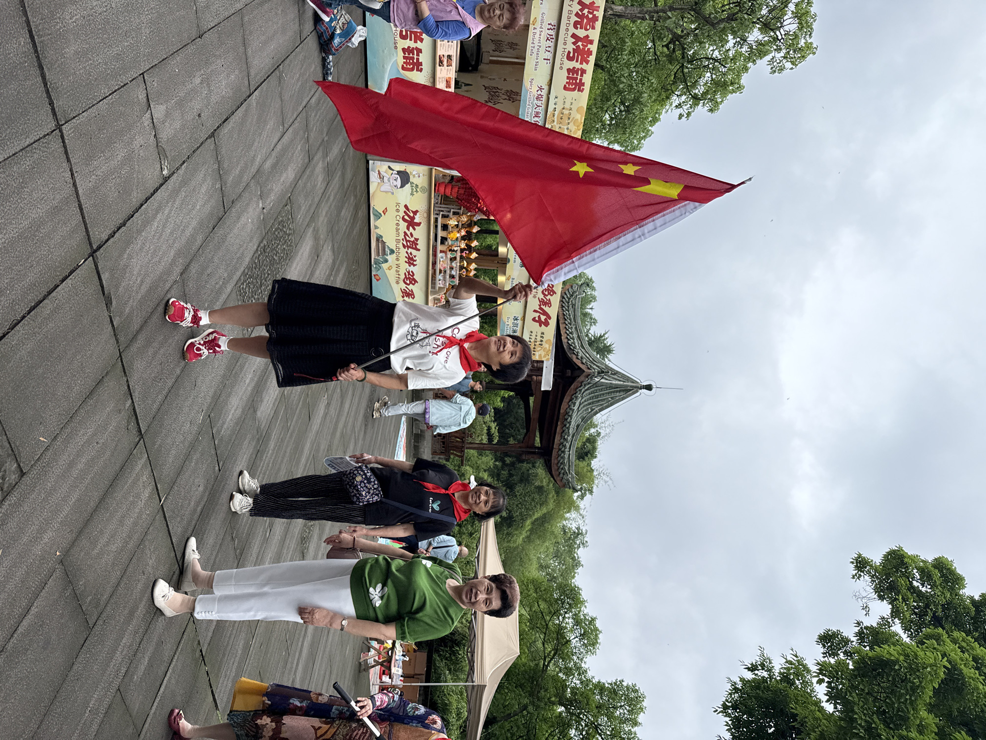 The image shows a woman standing on a brick walkway, holding a Chinese flag. She is wearing a white t-shirt with a red scarf around her neck, a black skirt, and red and white sneakers. Behind her are other people, including women wearing red scarves. In the background is a building with a traditional Chinese-style roof. There are also food stalls with signs in Chinese characters. The image was likely taken in a public space, perhaps a park or tourist area, in the Yuzhong District of China.