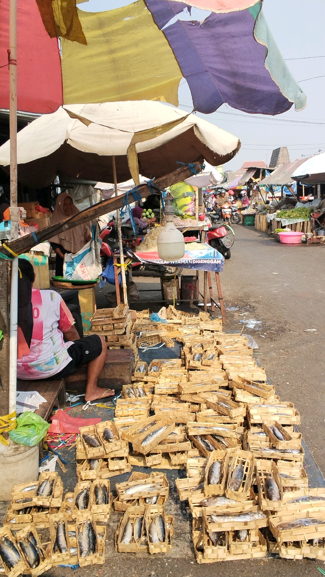 This is a street-level outdoor market scene in Mojokerto, Indonesia. The foreground is dominated by numerous small, woven bamboo baskets filled with fish, arranged in stacks on the ground. The baskets vary in size, and the fish appear to be dried or preserved. Behind the fish baskets, a person is seated on a wooden crate, facing away from the camera. They are wearing a white t-shirt with colorful graphics and black shorts.
Further back, a market stall is visible under a large, multi-colored awning made of fabric in shades of red, yellow, and purple. A woman wearing a headscarf is standing at the stall, which displays various goods including bags and what appears to be dried shrimp or small fish in baskets. A motorcycle is parked behind the stall.
In the background, the market continues with more stalls and shoppers. The ground is paved and appears to be a street or a designated market area. The overall impression is a busy, bustling marketplace selling fresh and preserved food items.