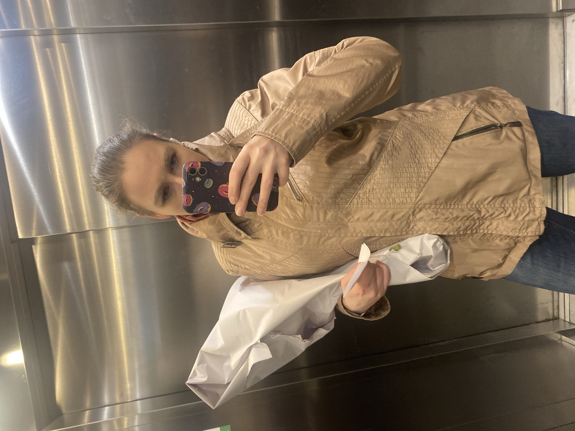 A person is taking a selfie in an elevator with a stainless steel interior. The individual is holding a phone in their right hand, which features a donut-patterned case. They are wearing a light brown jacket and blue jeans. They also hold a white bag in their left hand. The location cannot be determined from the image alone.