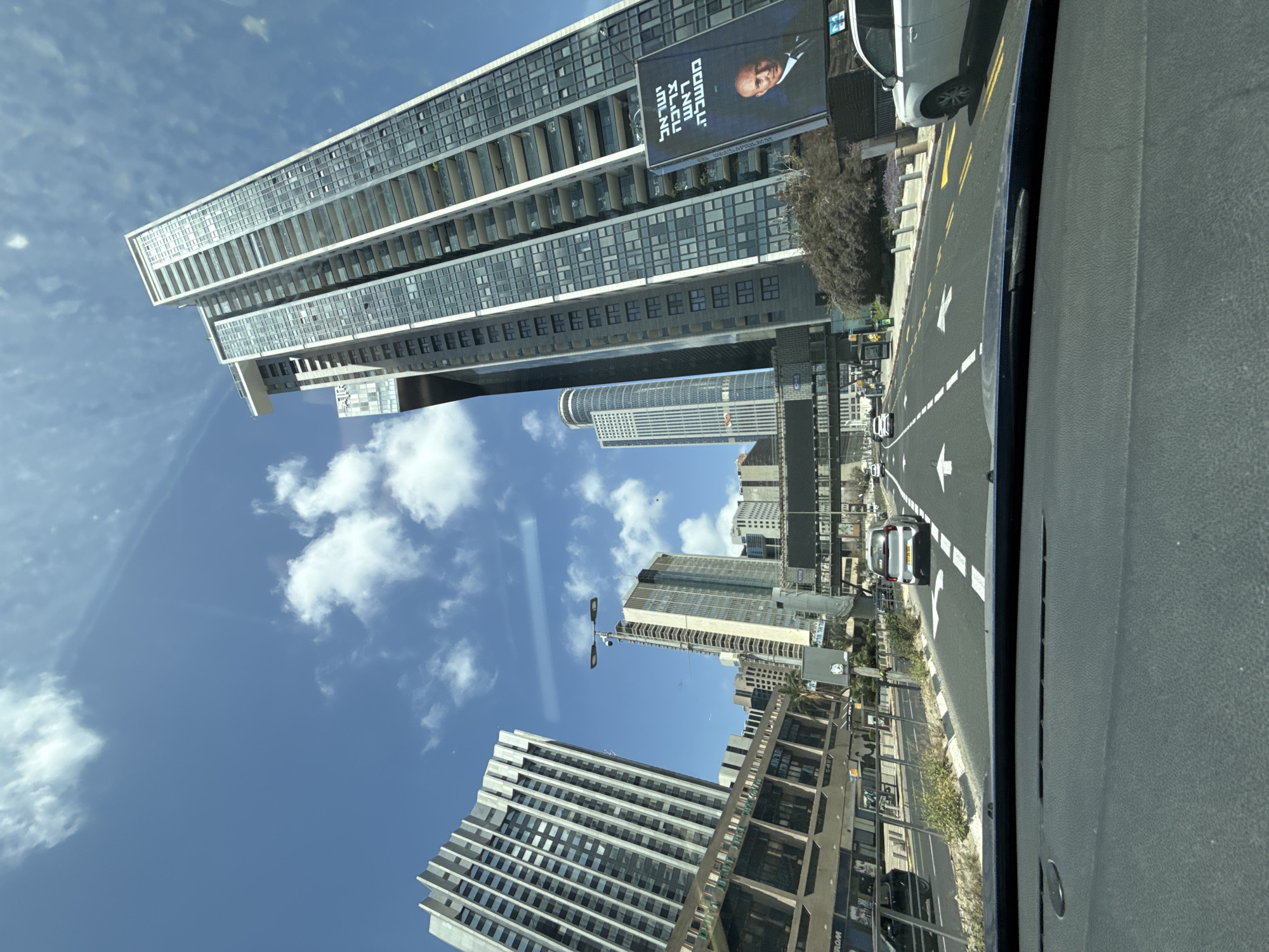 The image shows a view from inside a car, looking towards a street scene in Tel Aviv, Israel. The sky is visible with white clouds against a blue backdrop. The street includes multiple high-rise buildings, with at least five prominent buildings of varying heights and designs. A billboard with a portrait of a man is attached to one of the buildings. The street is marked with white directional arrows and yellow lines indicating traffic lanes. Cars and other vehicles are seen on the road. A section of the car's dashboard is visible on the right.