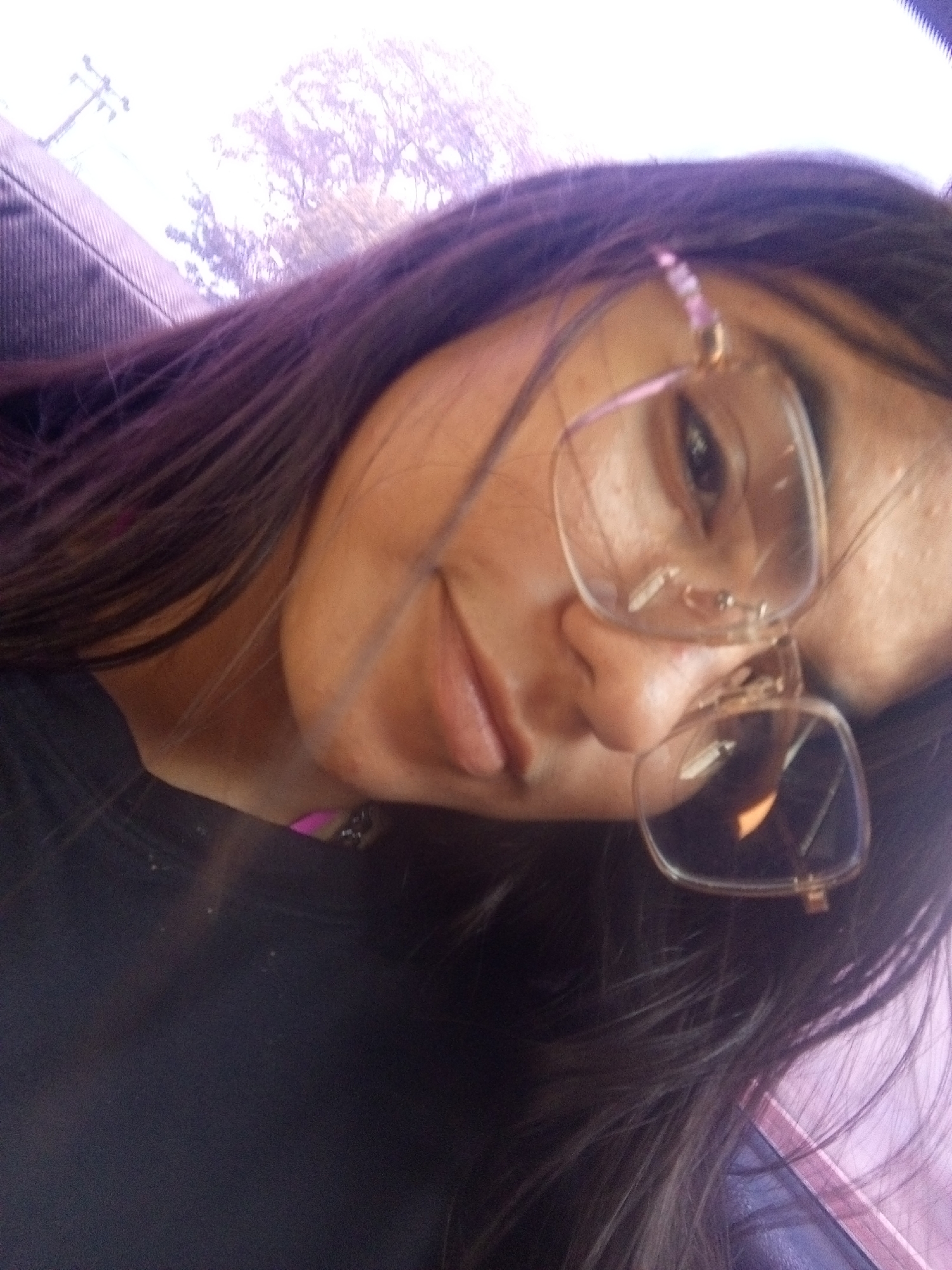 A close-up shot of a woman with long, dark hair wearing glasses. The photo is taken at an angle looking down towards the woman. Outside the car window there are trees, a building, and telephone poles. The location is within Juan Griego, Venezuela.