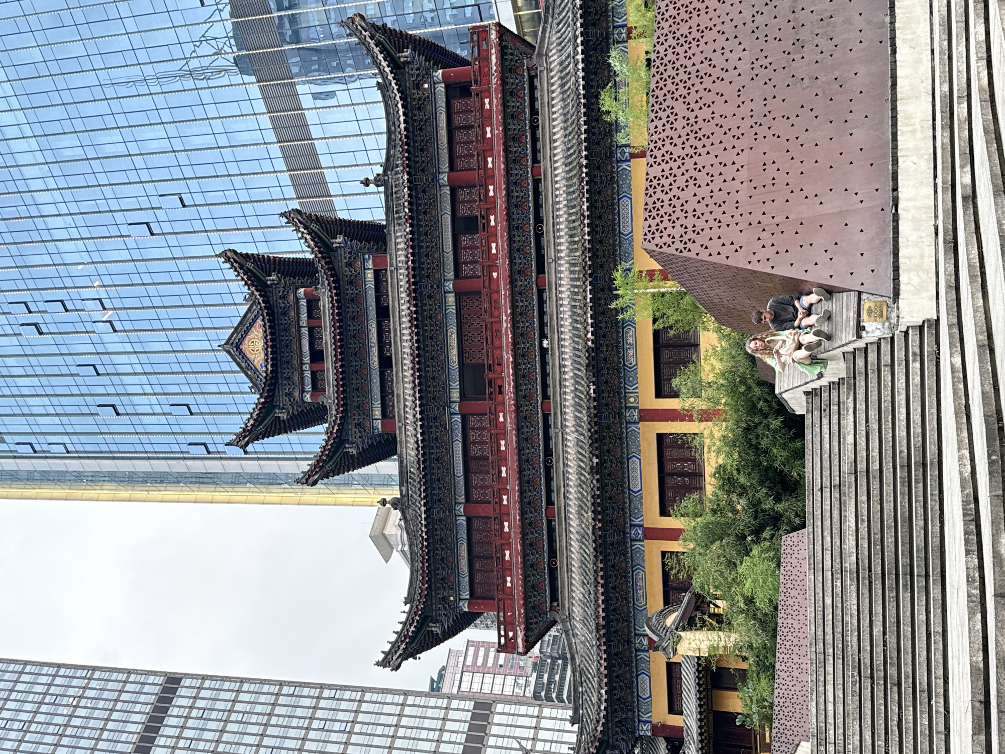 In Yuzhong District, China, a multi-tiered traditional Chinese temple with red and gold accents stands prominently in front of modern glass skyscrapers. Two individuals, a woman with blonde hair and a man with dark hair, are seated on a wide set of concrete stairs leading up to the temple. The woman is wearing a light green dress and carrying a tan bag, while the man is wearing a dark long-sleeved shirt and blue shorts. To their right is a large, rust-colored angular structure with a pattern of triangular perforations. Lush green bamboo and other foliage are visible between the temple and the modern structures, and on the steps. The sky is overcast.