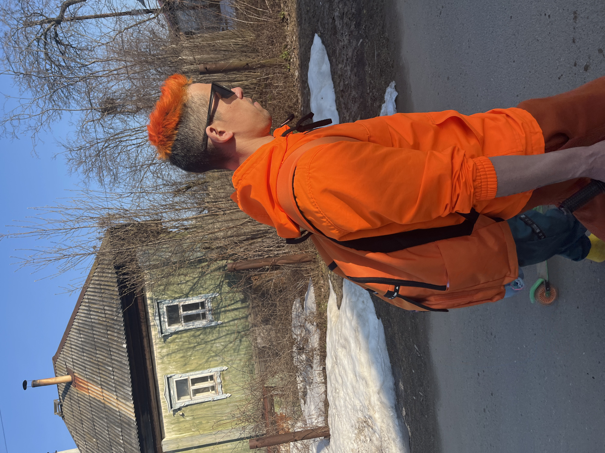 A man with orange hair and sunglasses is on a road next to a green wooden house with two visible windows. The man is wearing an orange jacket and has a backpack. A patch of snow is near the road. The location is in the city of Chaikovsky, Russia. The man is standing with a scooter to the side. The sky is blue and visible through bare branches.