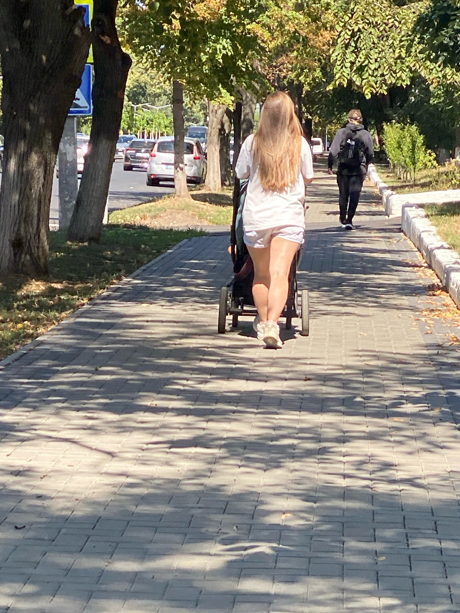 In the city of Chișinău, Moldova, a woman with long blonde hair, wearing a white t-shirt and white shorts, is pushing a stroller along a paved sidewalk lined with trees. The sun casts dappled shadows on the sidewalk from the leaves of the trees. In the background, a person wearing a dark hoodie and backpack walks away from the camera, also on the sidewalk. Further in the background, cars are visible on a street next to the sidewalk. The scene suggests a pleasant day in a park or a tree-lined residential area.