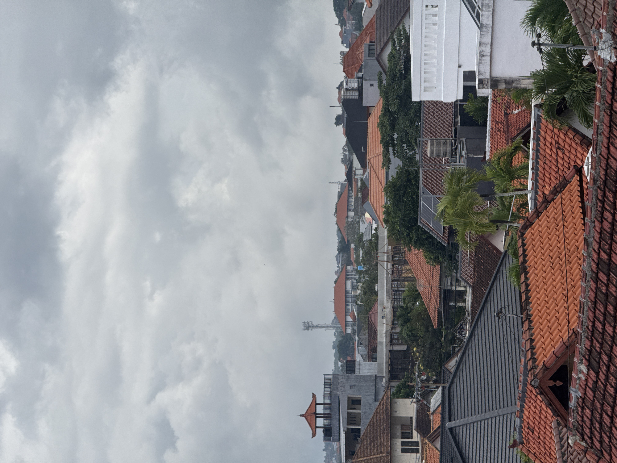 An overhead shot captures a dense collection of rooftops in a city, bathed in overcast, cloudy weather. The predominantly terracotta-tiled roofs are varied in shape and size, creating a textured mosaic across the frame. A few modern structures with flat roofs and white exteriors are interspersed among the traditional dwellings. Lush green trees peek out from behind and between the buildings, adding a natural element to the urban landscape. A tall communication tower is visible in the distance, hinting at the city's infrastructure. The scene is devoid of any visible human presence.