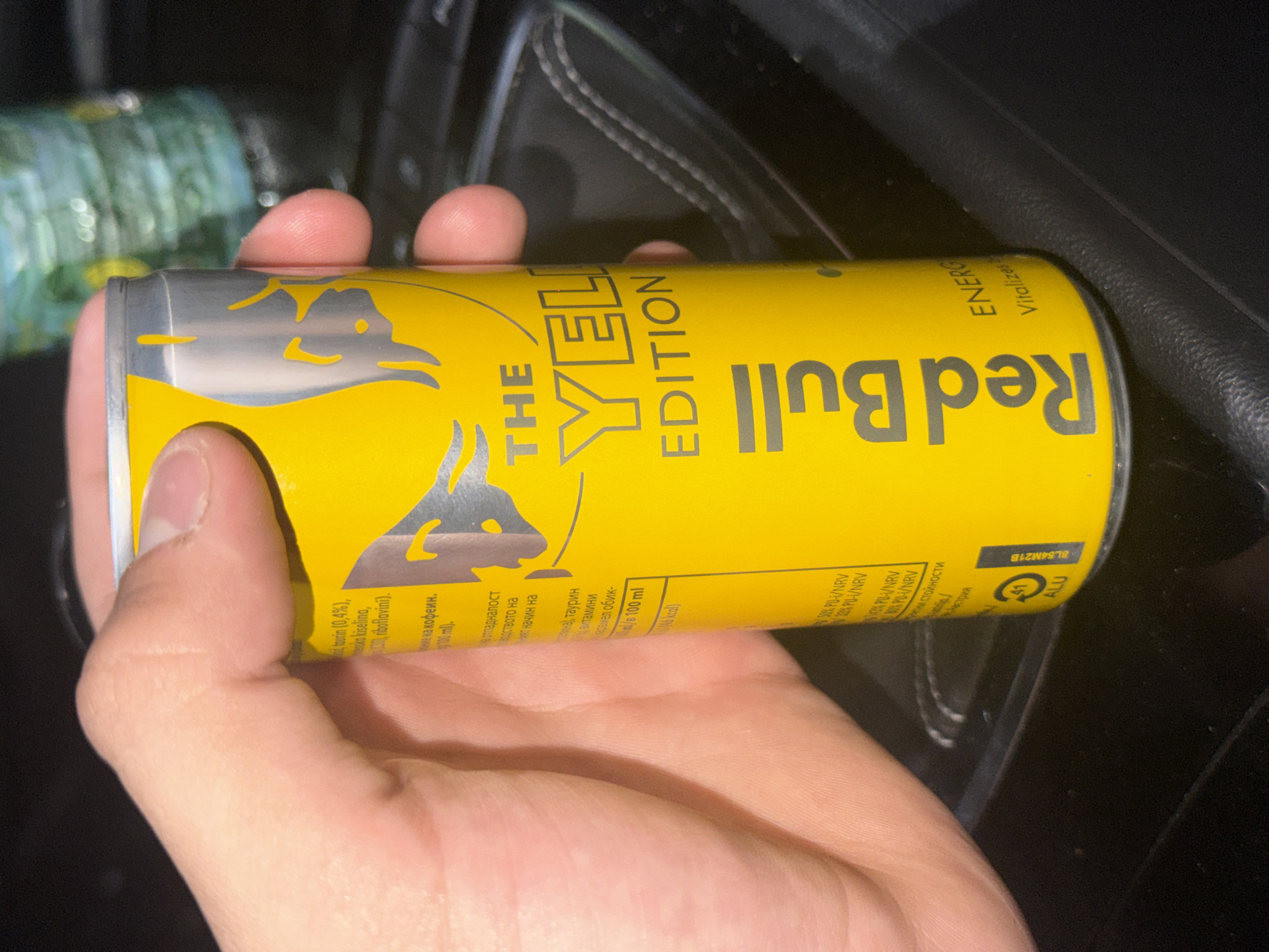An adult's right hand, characterized by fair skin, is depicted grasping a 250ml Red Bull energy drink can. The can is uniformly yellow with silver lettering, prominently displaying "THE YELLOW EDITION" and "Red Bull." Specific text visible on the can, written in Cyrillic, includes 'таурин (0,4%)' (taurine), 'киселина' (acid), 'рибофлавини' (riboflavin), 'кофеин' (caffeine), 'ml/в 100 ml', 'ккал' (kcal), and nutritional values such as '50% PU-I/NRV'. A recycling symbol indicating '41 ALU' (aluminum) and a production code 'BL54M21B' are present near the base, along with a reference to 'Австрия' (Austria). The hand is positioned over a dark, textured surface consistent with the interior of a vehicle, possibly a car console. A blurry, greenish-blue object is discernible in the distant background. Based on the Bulgarian language text on the Red Bull can, the scene is located in Varna, Bulgaria.