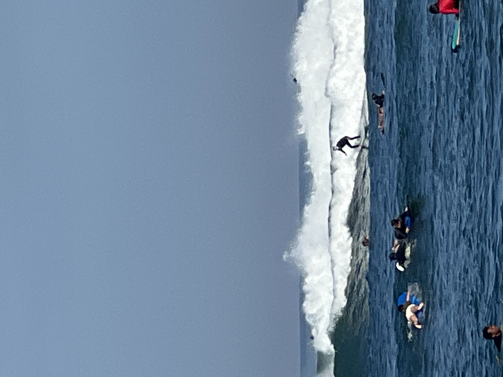This image depicts a coastal scene in North Kuta, Indonesia, with several surfers and individuals on surfboards in the ocean. The sky is clear and blue. The water is a deep blue, with white waves breaking in the middle ground.
In the middle of the image, a surfer in a black wetsuit is riding a wave. To the left of this surfer, a group of people are lying on surfboards, some appearing to be children, one wearing a light-colored swimsuit. To the right of the main surfer, another surfer is lying on a board. Further in the background, on the breaking waves, more surfers are visible. In the foreground, a person in a red top is sitting on a surfboard. The overall environment suggests a sunny day at a popular surfing spot.