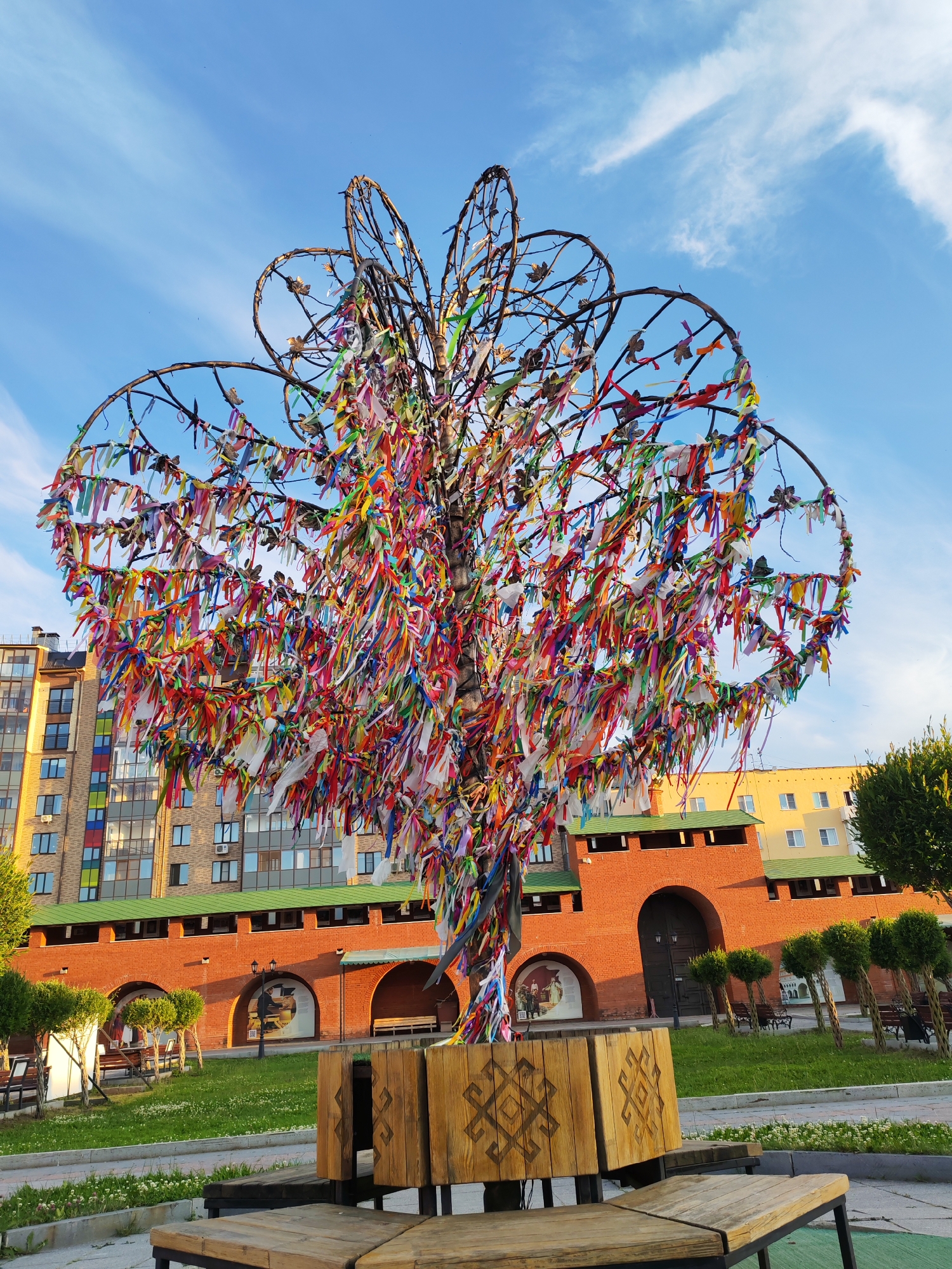 The image shows a large, artificial tree adorned with numerous colorful ribbons. The tree has a metal frame shaped like a stylized tree with large, circular branches. It stands in an outdoor public space in Yoshkar-Ola, Russia. In the background, there is a modern apartment building with a multi-colored facade, and a historic-looking red brick building with arched doorways and wooden gates. The ground in front of the brick building is a manicured lawn with several small, manicured trees and benches. The sky is blue with some wispy clouds. The image is taken from a low angle, looking up at the tree and the surrounding architecture. There are no people visible in the image.